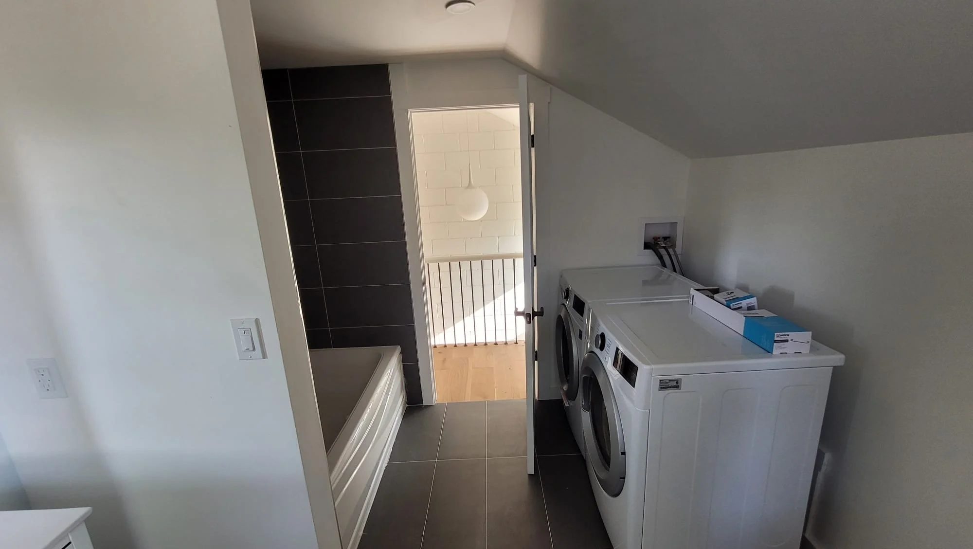 Laundry room with white washing machine and dryer, dark gray tiled floor, and a door leading to a balcony with a white railing and a round pendant light.