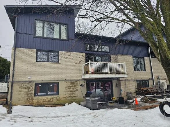 A multi-story house with beige brick lower half and blue upper siding, showing multiple windows, a small balcony, and snow on the ground with some construction or yard work tools outside.