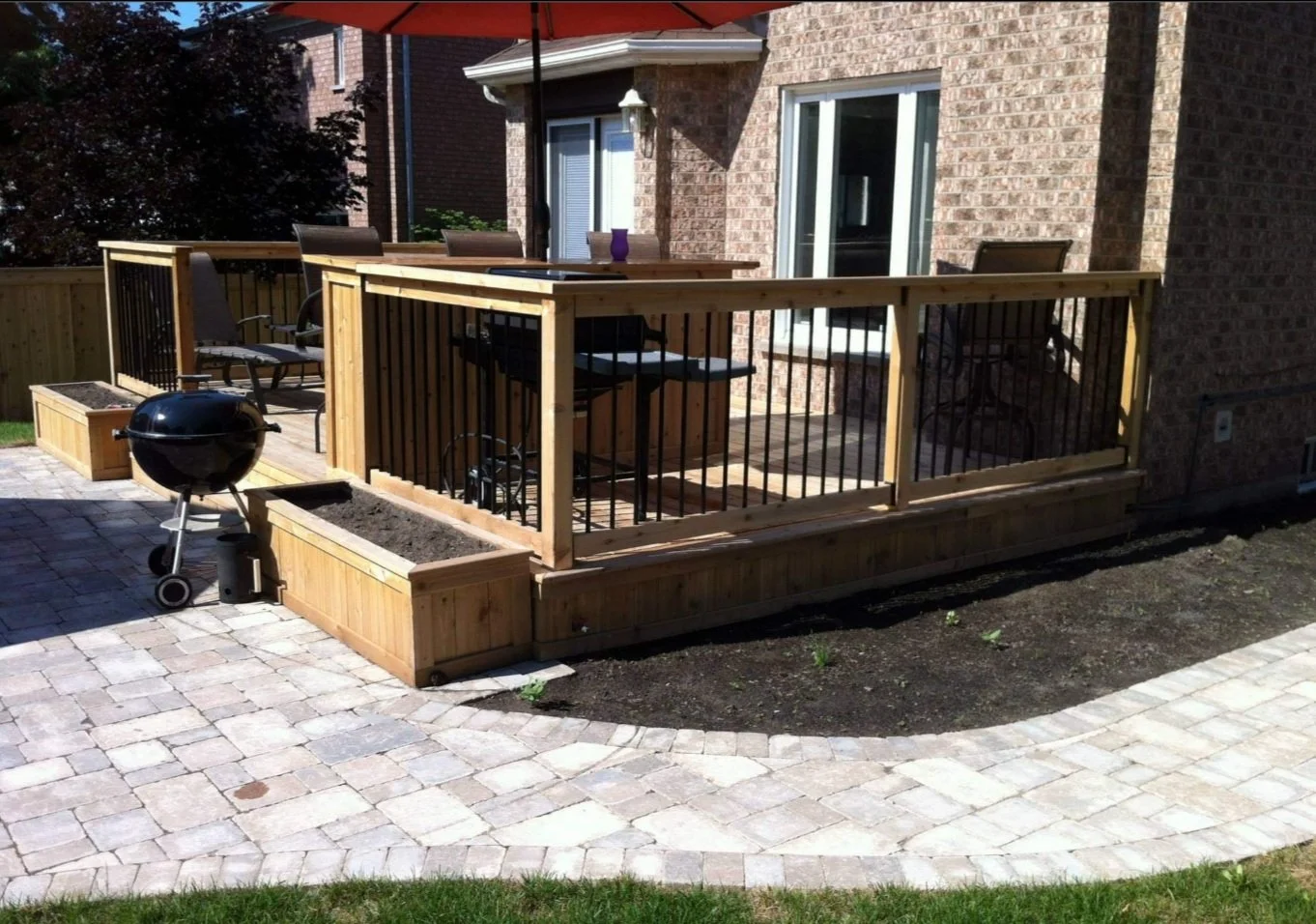 A newly built wooden deck with black metal railing attached to a brick house. The deck has outdoor furniture, including chairs and a table, and a small barbecue grill nearby. There are raised garden beds along the edge of the deck, with some soil ins
