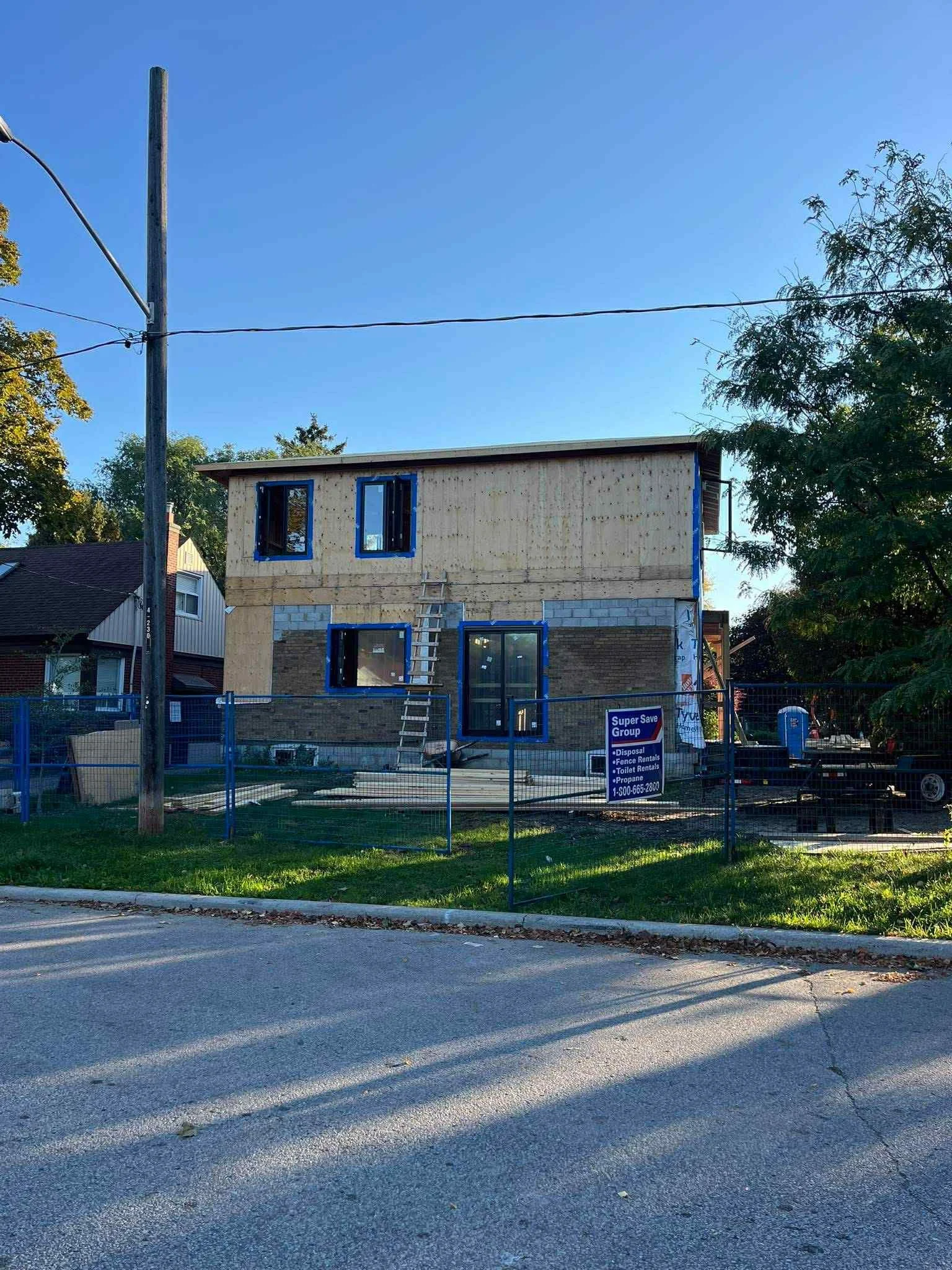 A two-story house under construction with wooden and brick exterior walls, several open window frames, and a ladder leaning against the house. A construction sign is visible on the fence in front of the house, and there are trees, a utility pole, and