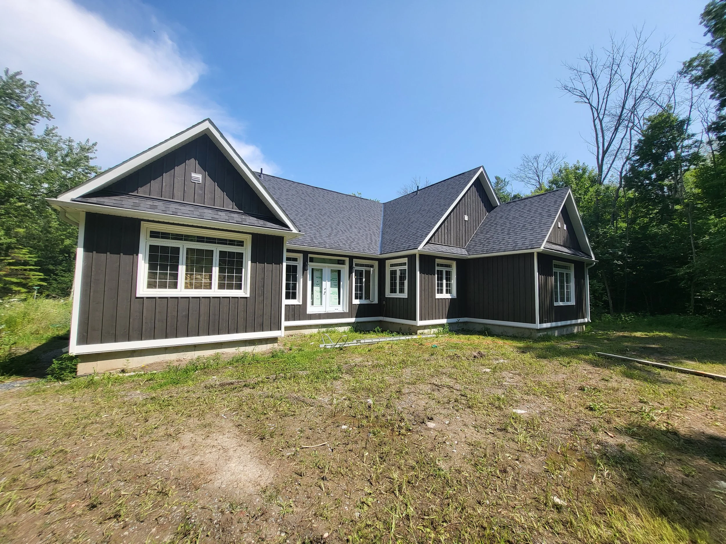 Newly constructed black house with multiple steep gabled roofs, white window frames, and a partially finished yard, surrounded by trees under a blue sky.