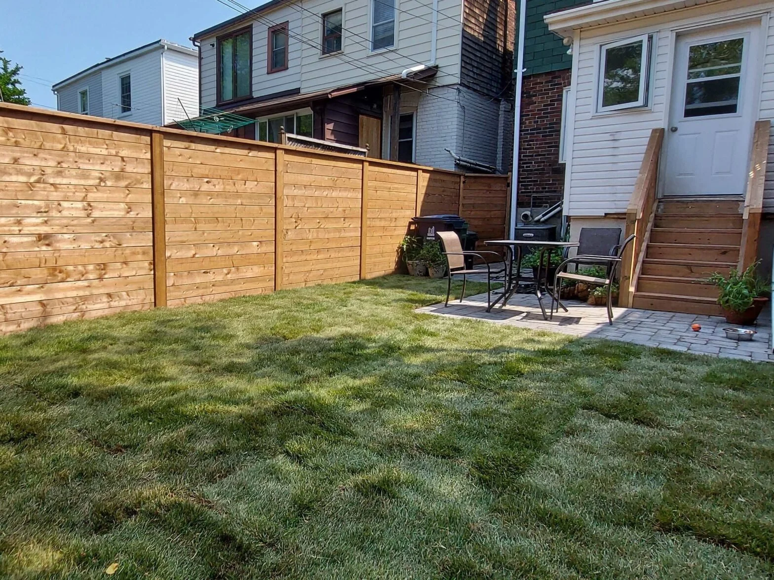 Backyard with a green lawn, wooden privacy fence, patio area with outdoor table and chairs, potted plants, and stairs leading to a door.