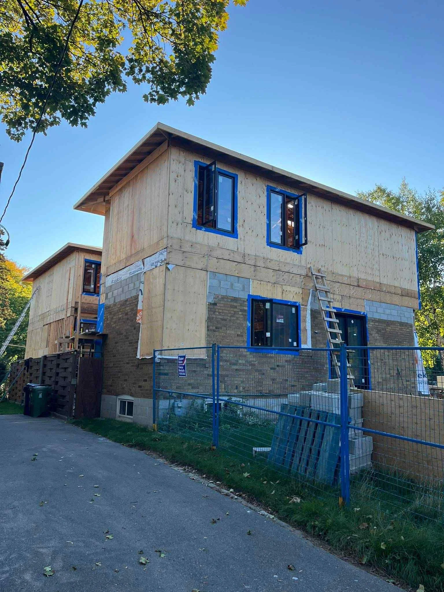 A two-story house under construction with the lower part made of brick and the upper part covered in wooden panels. Several open window frames are visible, and a wooden ladder leans against the house. There is a blue fence surrounding the constructio