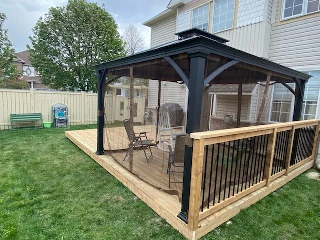 A screened-in backyard patio with wooden flooring, outdoor furniture, and a black and beige railing, attached to a beige house with large windows.
