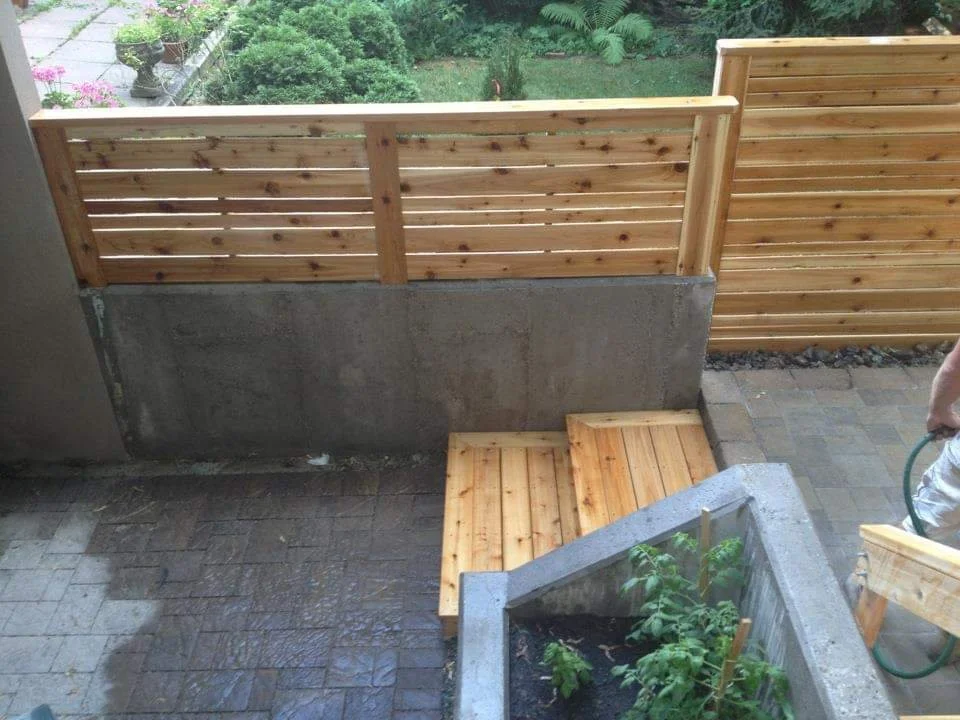View of a backyard patio with newly installed wooden fencing and a small wooden decked platform, with a garden hose and hand of a person visible.