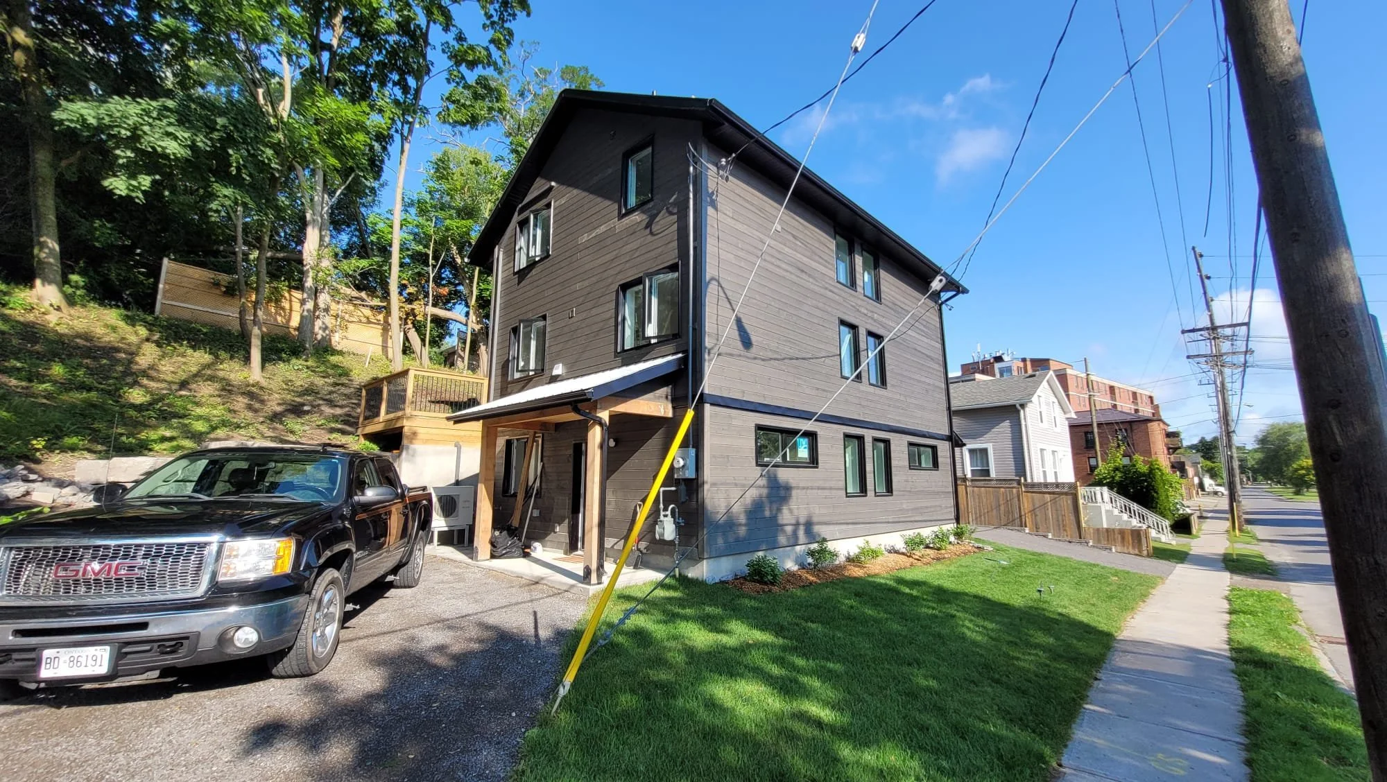 A three-story black residential building with a small porch and green lawn in a neighborhood. A black GMC truck is parked in front. The background features other houses, a sidewalk, trees, utility poles, and a blue sky with some clouds.