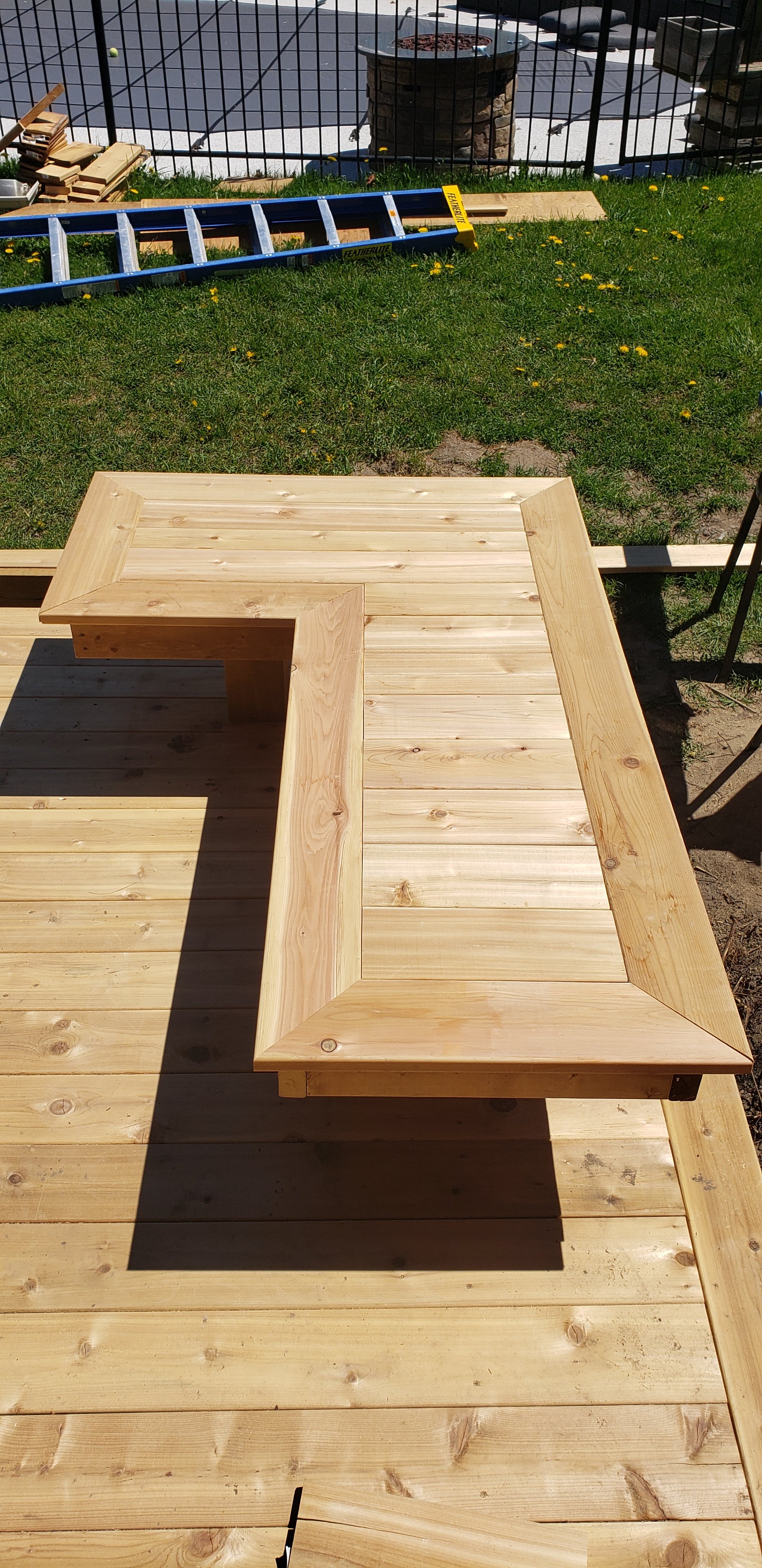 A wooden deck with an L-shaped design, surrounded by grass, during daytime. Construction tools and materials, including a blue ladder, are visible in the background.