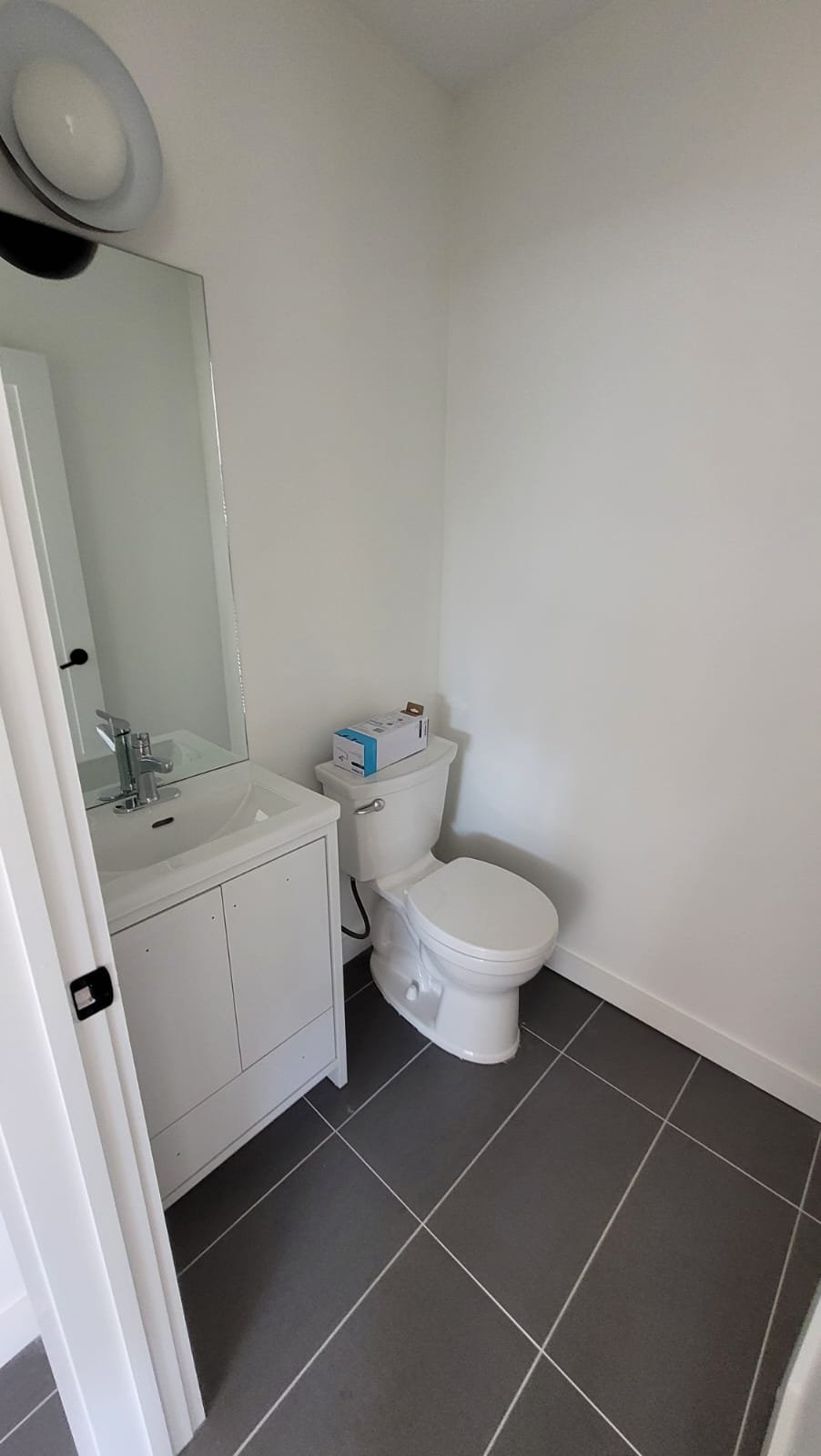 A small bathroom with gray tiled floor, a white toilet with a box on top, a white vanity with a sink, and a large mirror above the sink, with plain white walls.