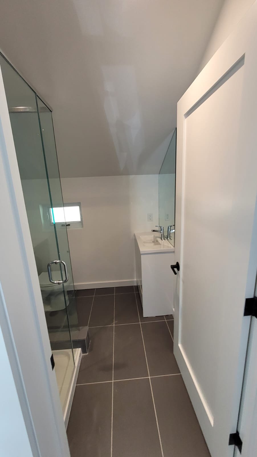 Small bathroom with a glass shower stall, a white vanity with a sink, and a tiny window on the wall. The floor has large dark gray tiles, and the room has white walls and ceiling.