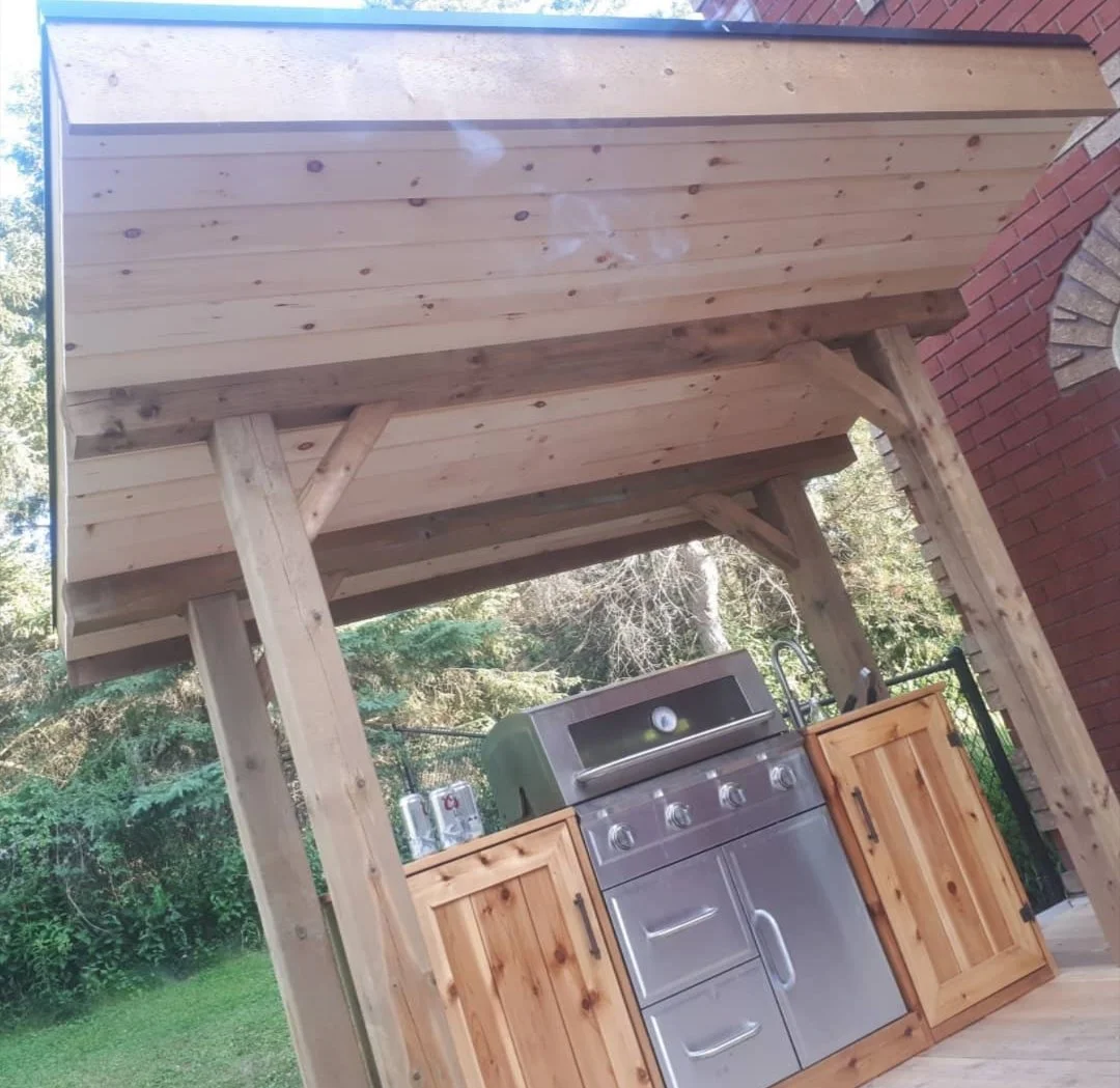 Outdoor kitchen with stainless steel grill, wooden cabinets, and a wooden roof structure in a backyard with trees in the background.