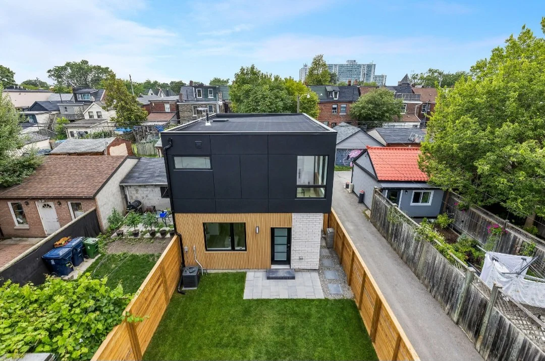A modern two-story house with black and wooden exterior panels, a small backyard with a grassy lawn, and neighboring houses with trees in a suburban neighborhood.