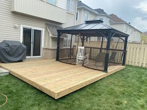 Backyard wooden deck with a gazebo, outdoor furniture, and a grill, surrounded by a light-colored house and a wooden fence.