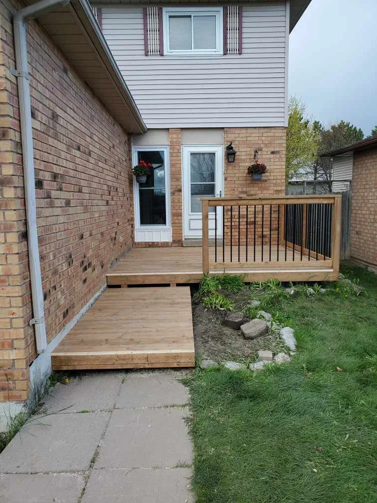 View of a small wooden deck and ramp attached to a house, with brick and siding exterior, a window with flower boxes, a door, and a small garden area with rocks and plants.