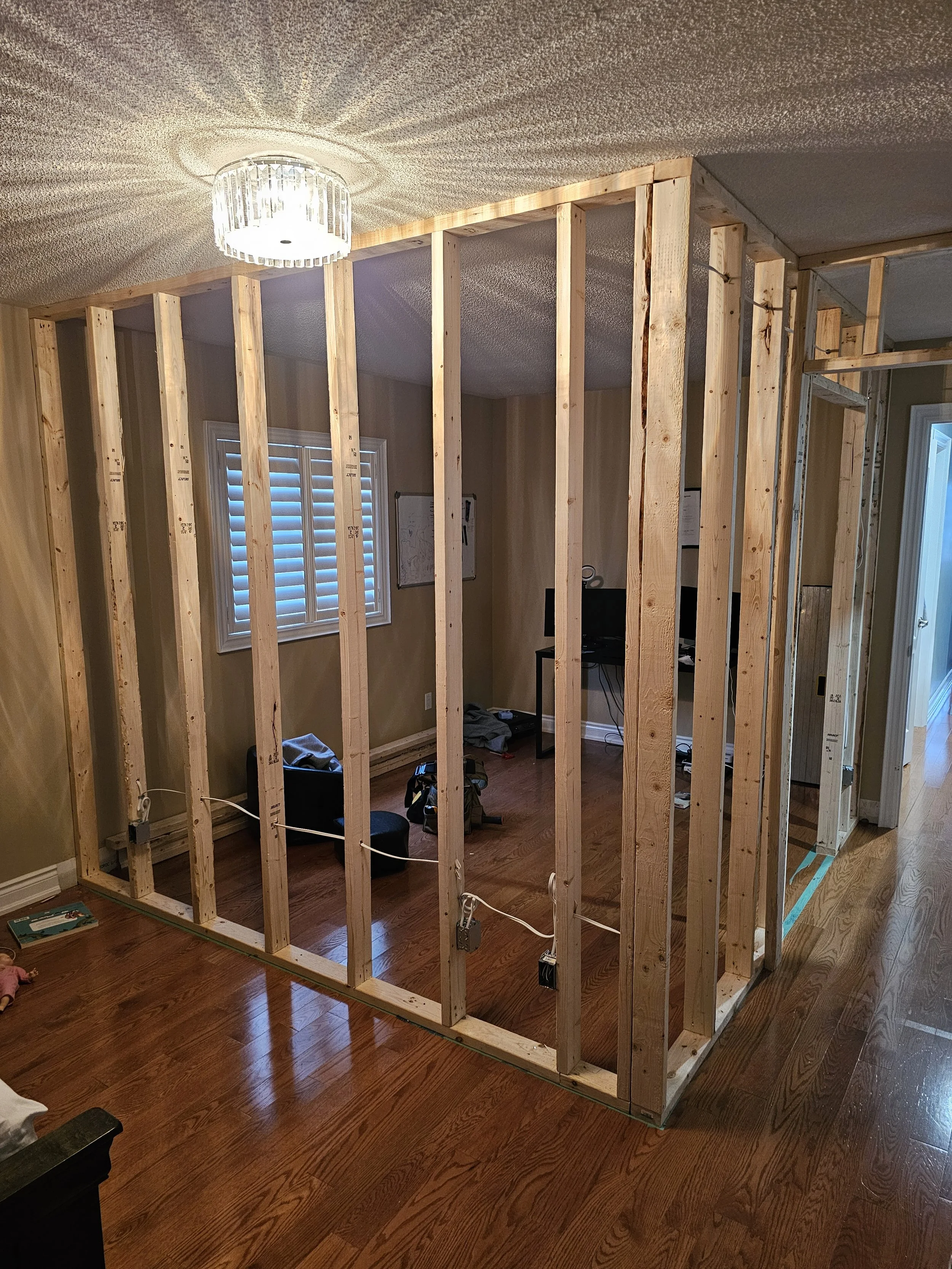 Wooden framing of a new wall or room being built inside a house with hardwood floors, a window with blinds, and a ceiling light fixture.