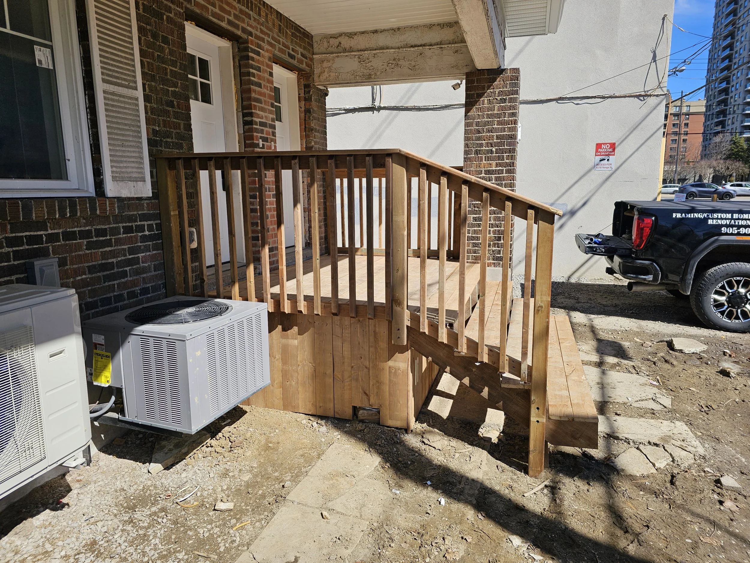 New wooden porch with stairs and railing outside a brick building, with HVAC units nearby and a black pickup truck parked in the background.