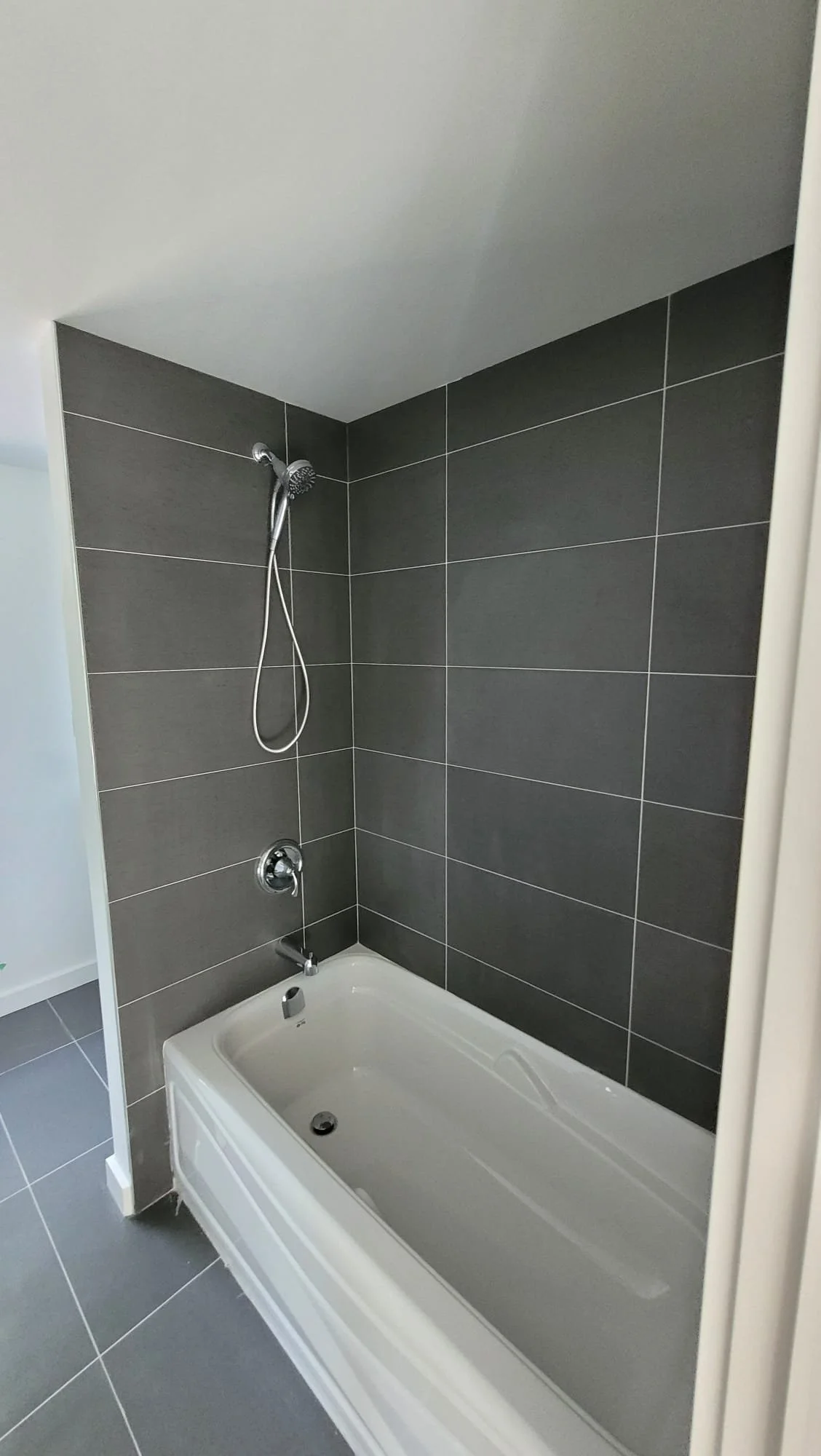 A bathroom shower with a white bathtub, dark gray tiled walls, and a silver showerhead with a flexible hose.
