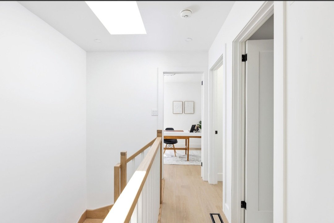 Bright, minimalist hallway with white walls, light wood flooring, and a stair railing on the left. At the end of the hall, a small workspace with a desk, chairs, and wall art is visible.