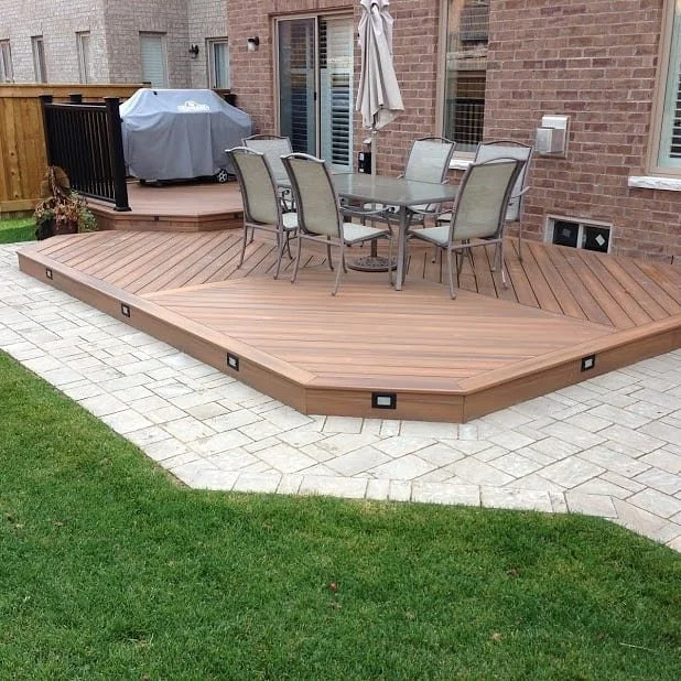 A wooden raised deck with a glass-top table and six chairs, a grill covered with a tarp, and a brick house in the background.