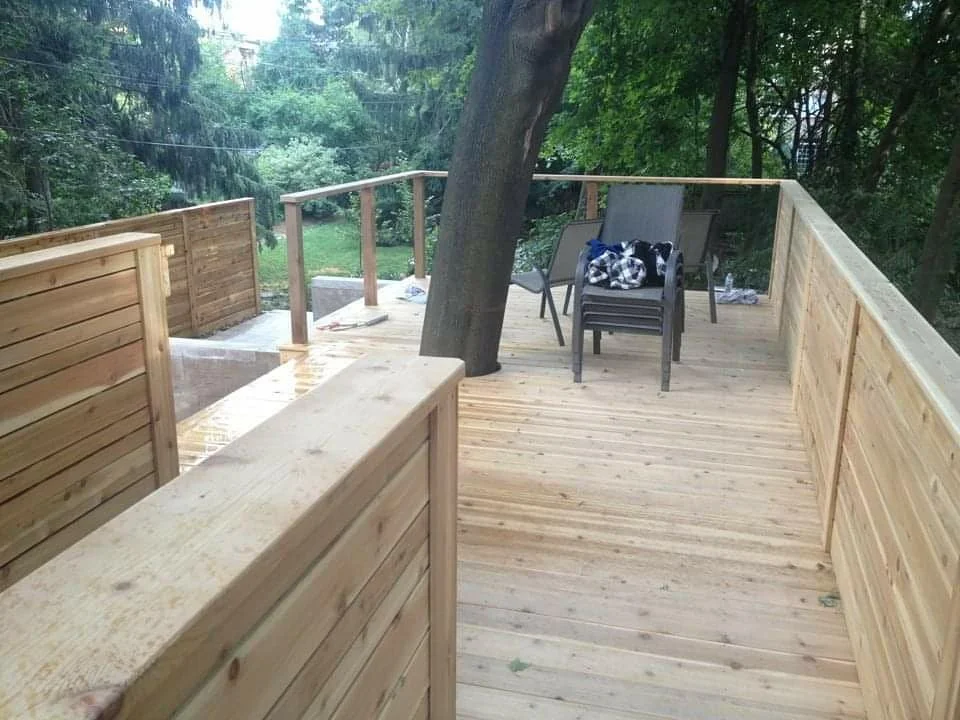 A newly built wooden deck with railing, positioned around a tree in a wooded backyard. There are a few chairs and a cloth on one of them, with some tools and construction materials on the deck.