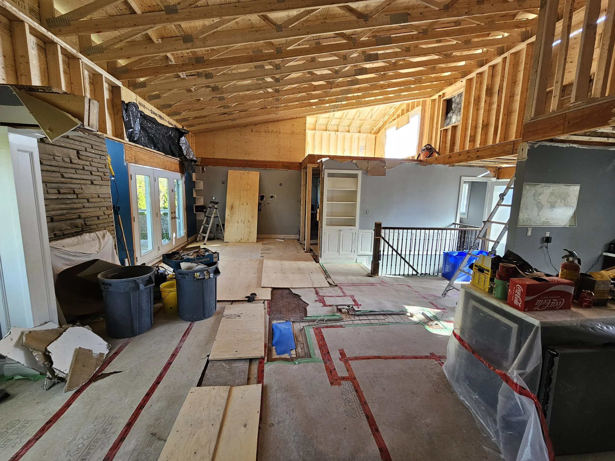 Interior of a house under construction with exposed wooden beams, partially installed flooring, construction tools and materials, ladders, and an open staircase railing.