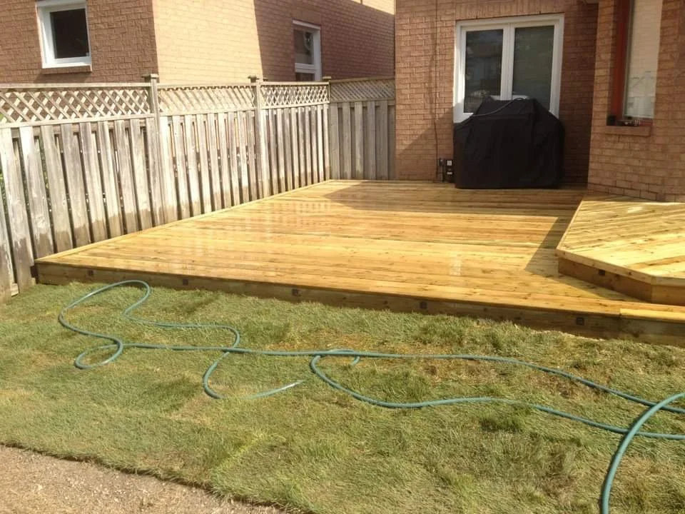 Freshly constructed wooden deck in a backyard, with garden hose on the grass, a black grill cover, and a brick house in the background.
