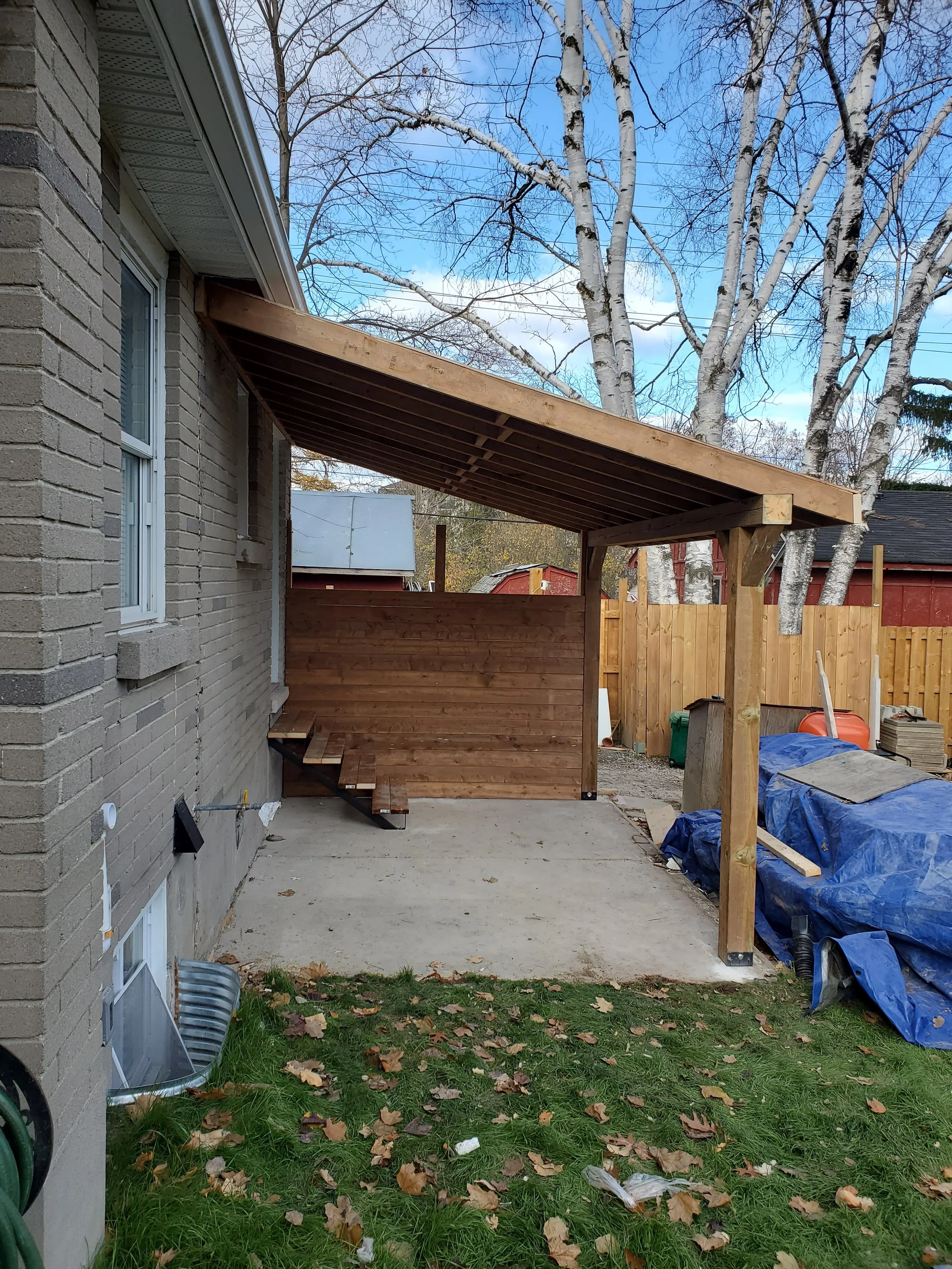 Construction of a wooden porch and privacy wall outside a house with a concrete patio, fallen leaves, and trees in the background.