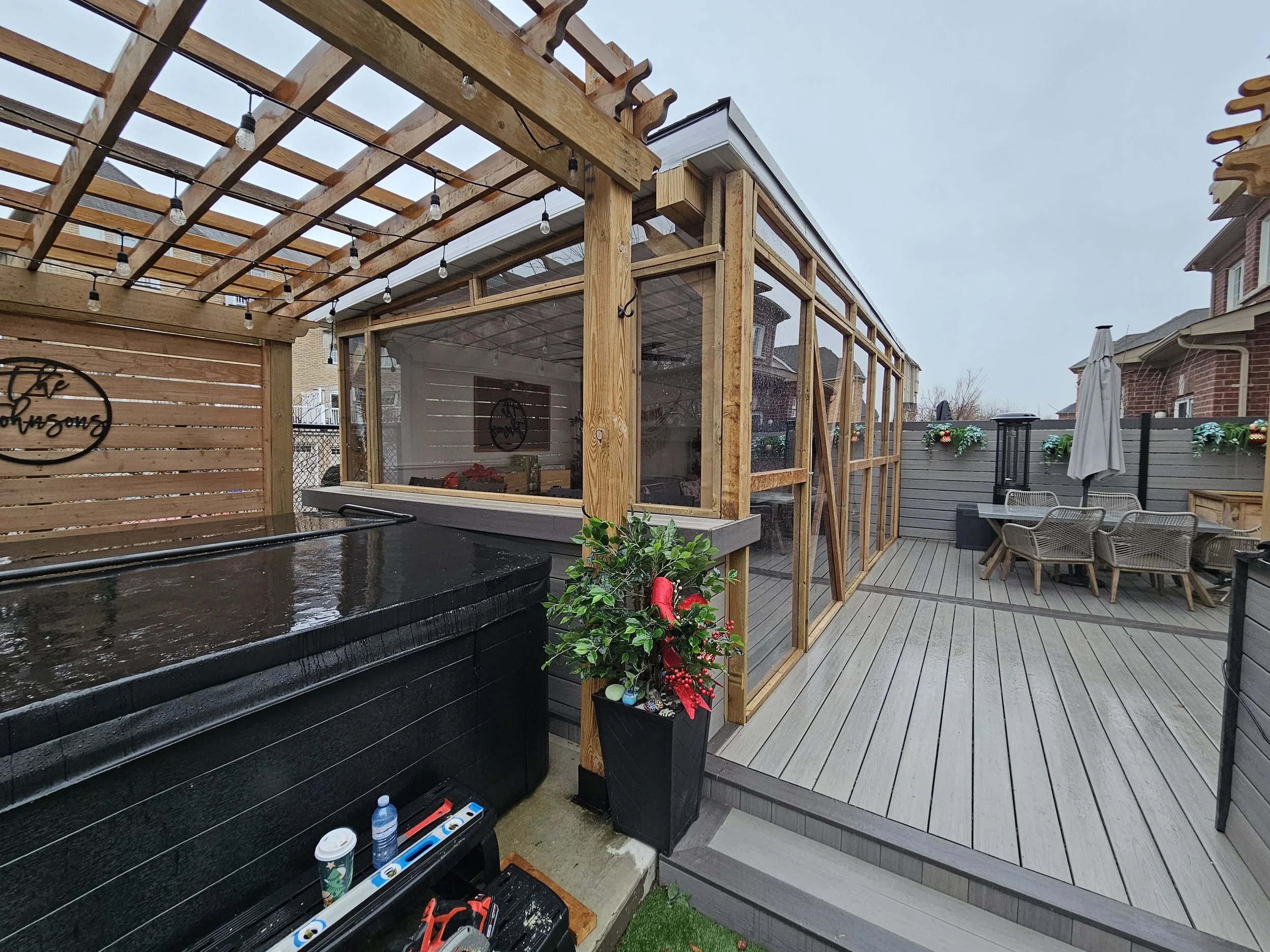 A backyard patio with a hot tub, wooden fence with decorative sign, potted plant, and a weatherproof enclosure with transparent walls covering part of the deck. There's outdoor seating with a table, chairs, and an umbrella, along with festive decorat