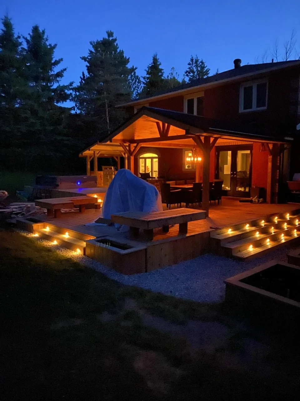 A house with a wooden deck illuminated by string lights at dusk, surrounded by trees.