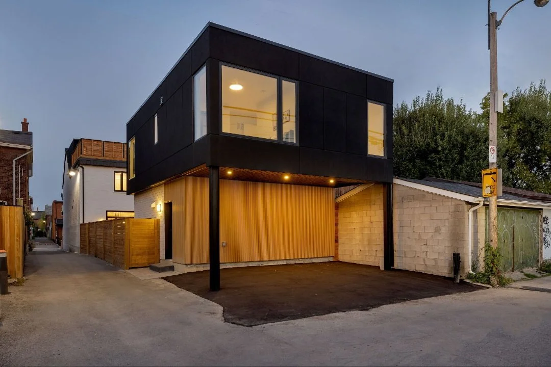 Modern two-story building with black and wooden exterior, large windows, and an open space below supported by a black pillar, situated on a street with neighboring houses and a utility pole.