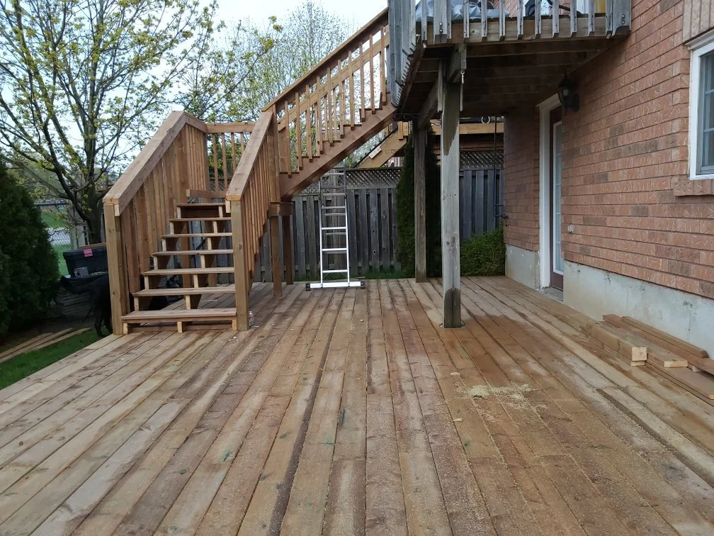 A wooden deck under construction with stairs leading up to an elevated balcony, next to a brick house, with a ladder, some scattered wood blocks, and trees in the background.