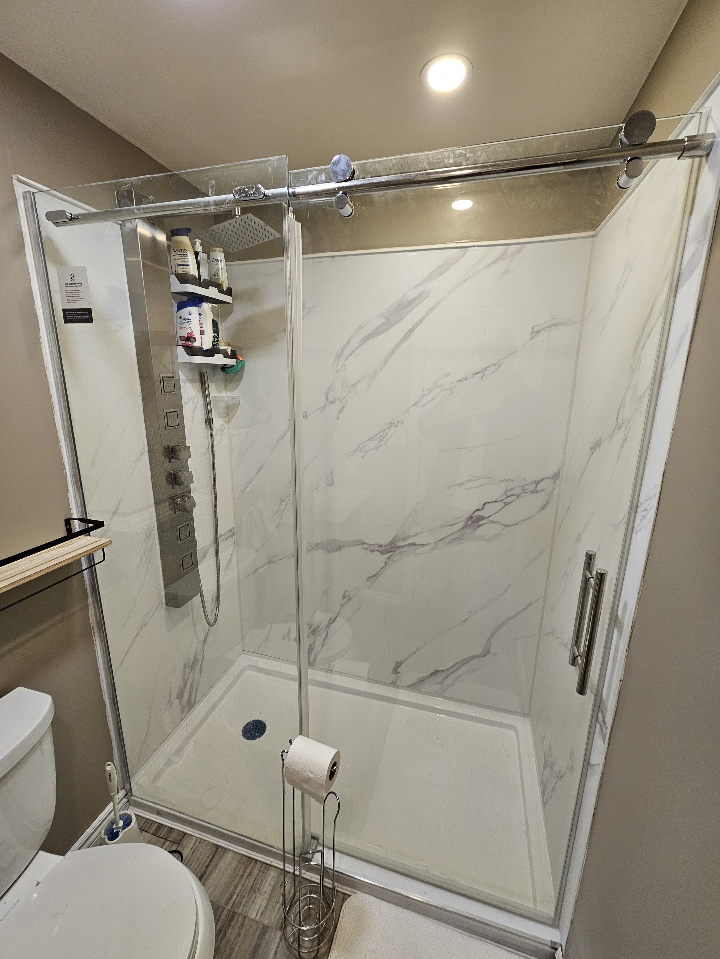 A modern walk-in shower with marble walls, a glass door, and a shower panel with multiple controls and shower heads. There are bottles of toiletries on a shelf inside the shower. A toilet is visible in the foreground, and a toilet paper holder with a