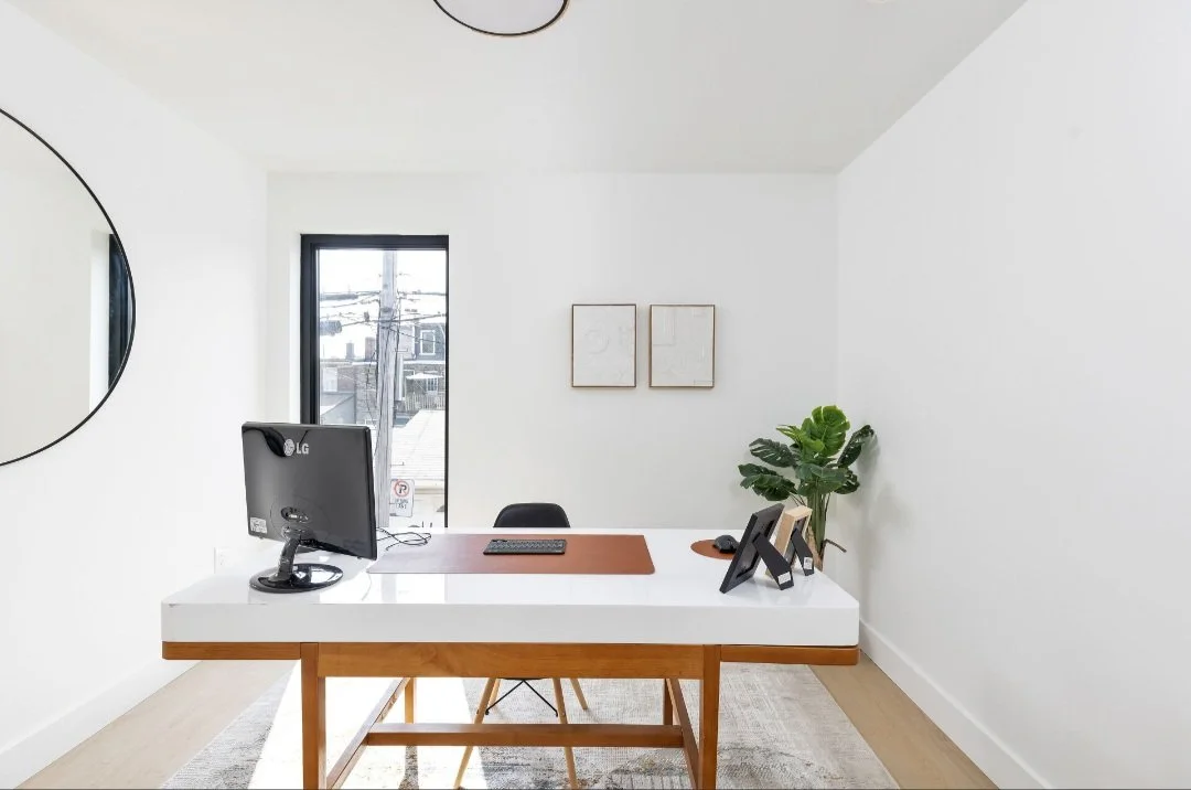 Modern office space with a white desk, computer monitor, framed pictures, potted plant, and minimalist decor.