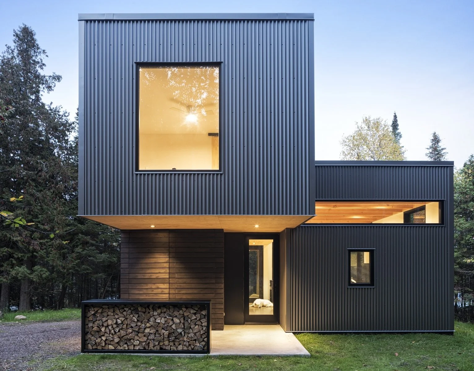 Modern black house with large windows and wood accents, surrounded by trees at dusk.