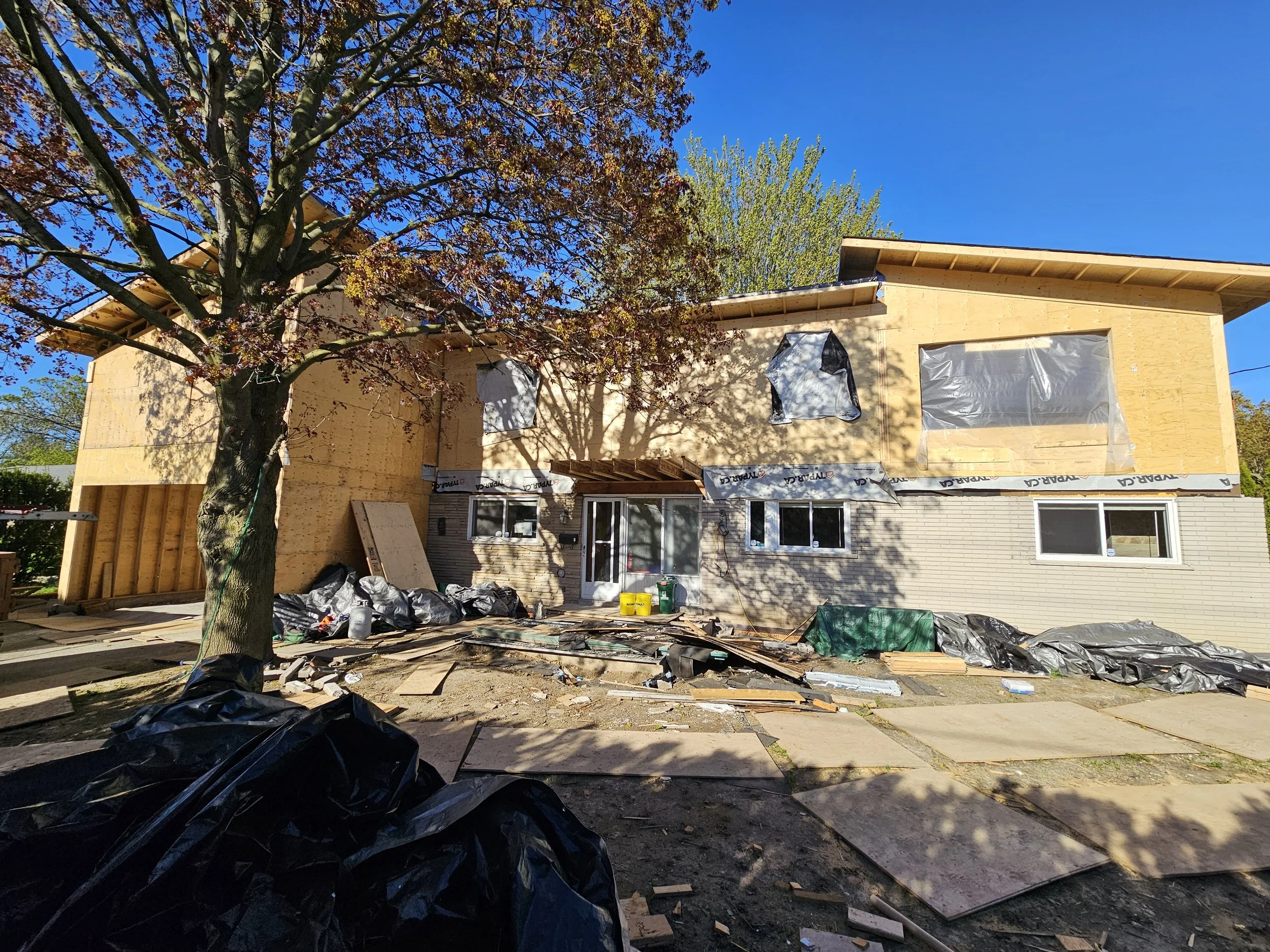 A house under construction with scaffolding and building materials, a large tree in the front yard, and a bright blue sky.