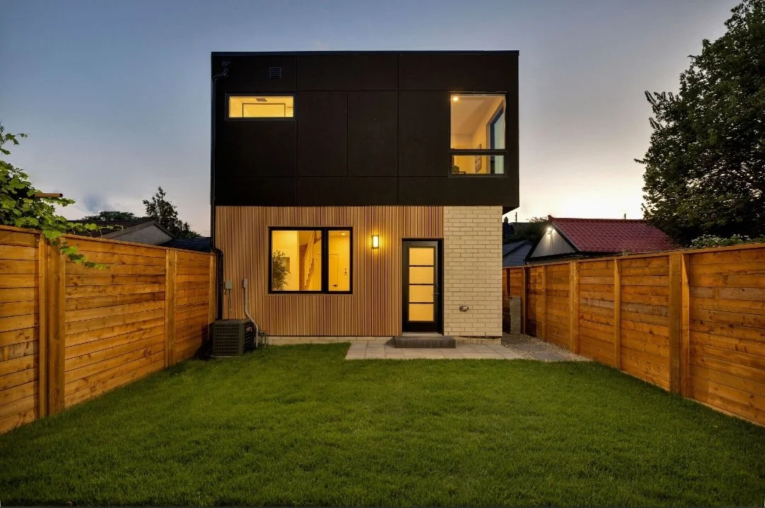 Modern two-story house with black upper and mixed wood and brick lower exterior, illuminated windows and door, fenced backyard with grass, trees, and neighboring rooftops at dusk.