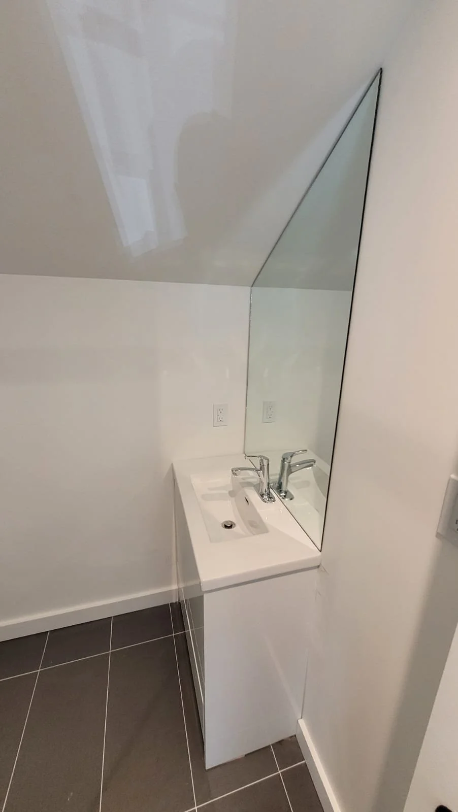 A small bathroom vanity with a white cabinet, a white sink, chrome faucet, and a large frameless mirror on the wall behind, with two electrical outlets nearby.