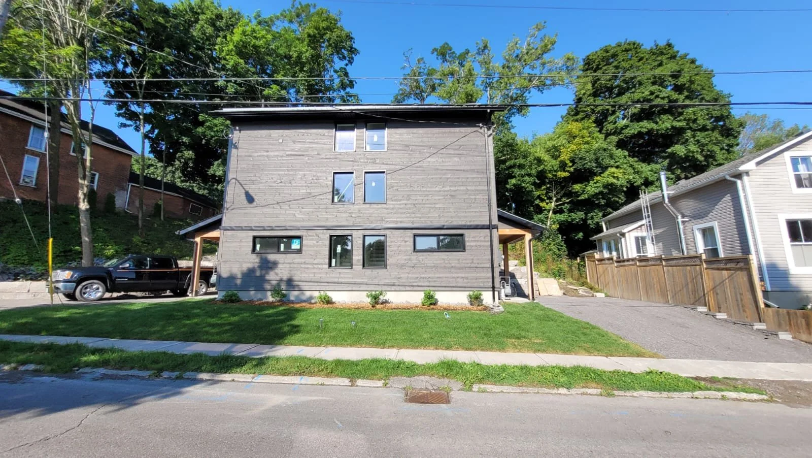 Front view of a gray modern residential building with a small landscaped lawn and a paved driveway on the right, surrounded by trees and neighboring houses under a clear blue sky.