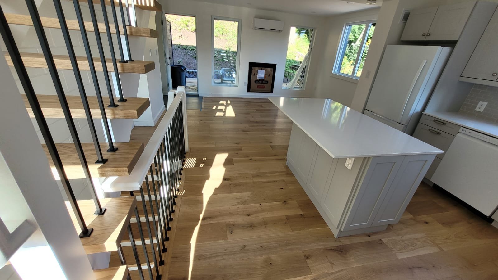 Interior of a modern kitchen and dining area with wooden floors, white cabinets, an island with a white countertop, and a staircase with wooden steps and black metal railing.
