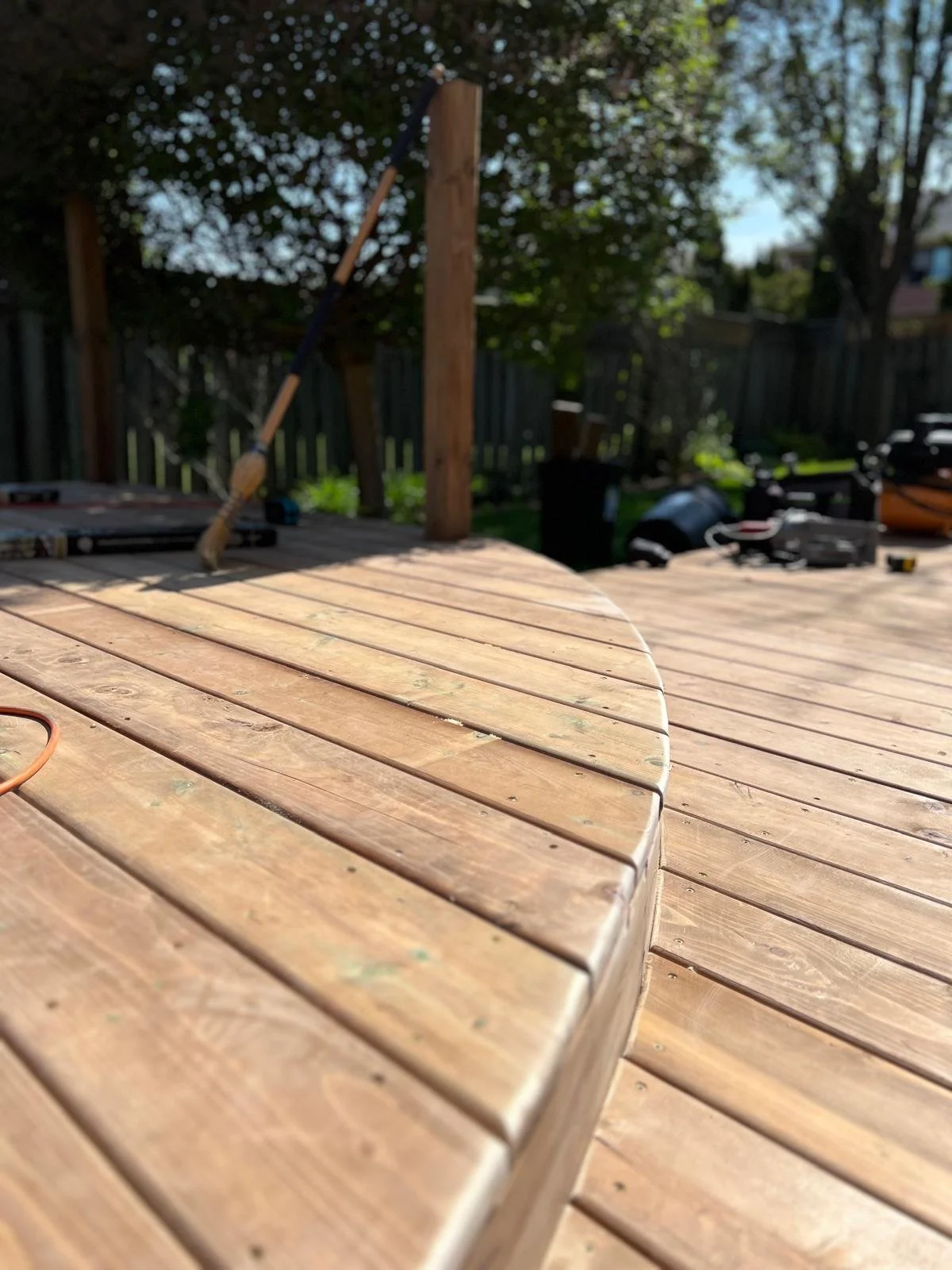 Close-up of a newly stained wooden outdoor deck with construction tools and supplies in the background on a sunny day.