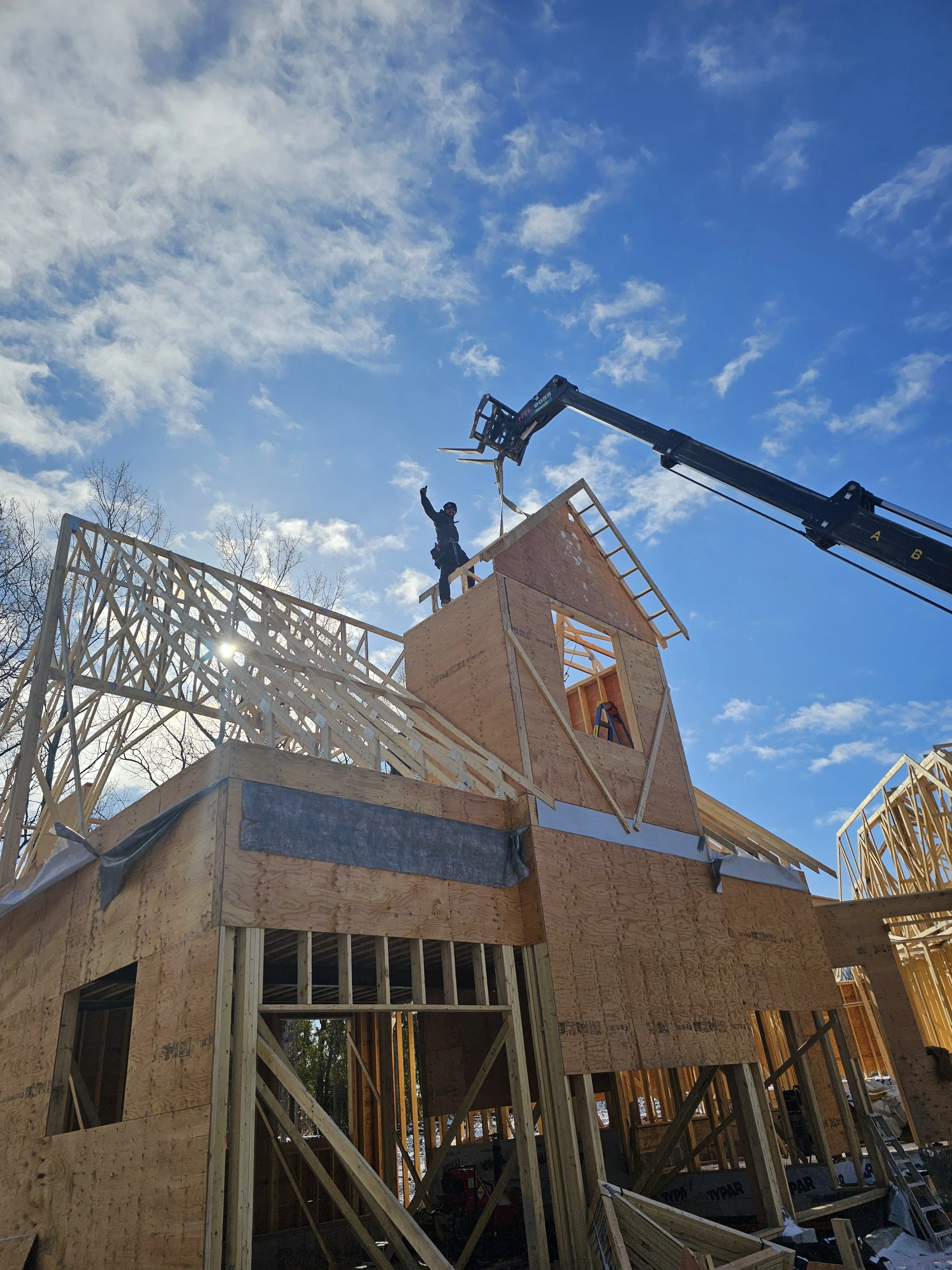 A person standing on the roof of a house under construction, waving, with a crane lifting a wooden beam against a blue sky with some clouds.