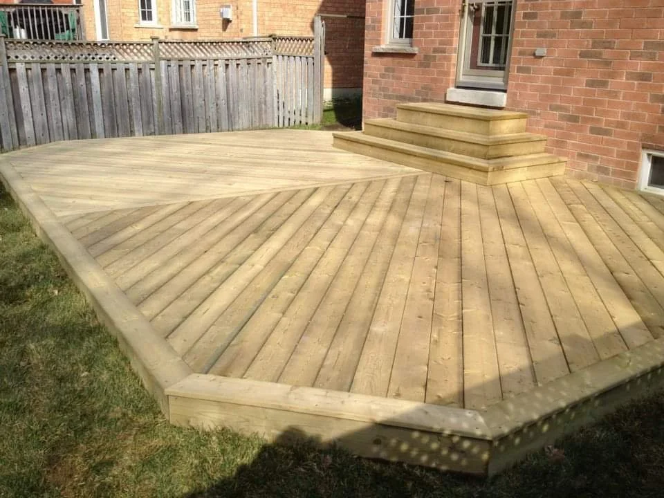 A newly built wooden deck attached to a brick house, with steps leading to a door, and a fence in the background.