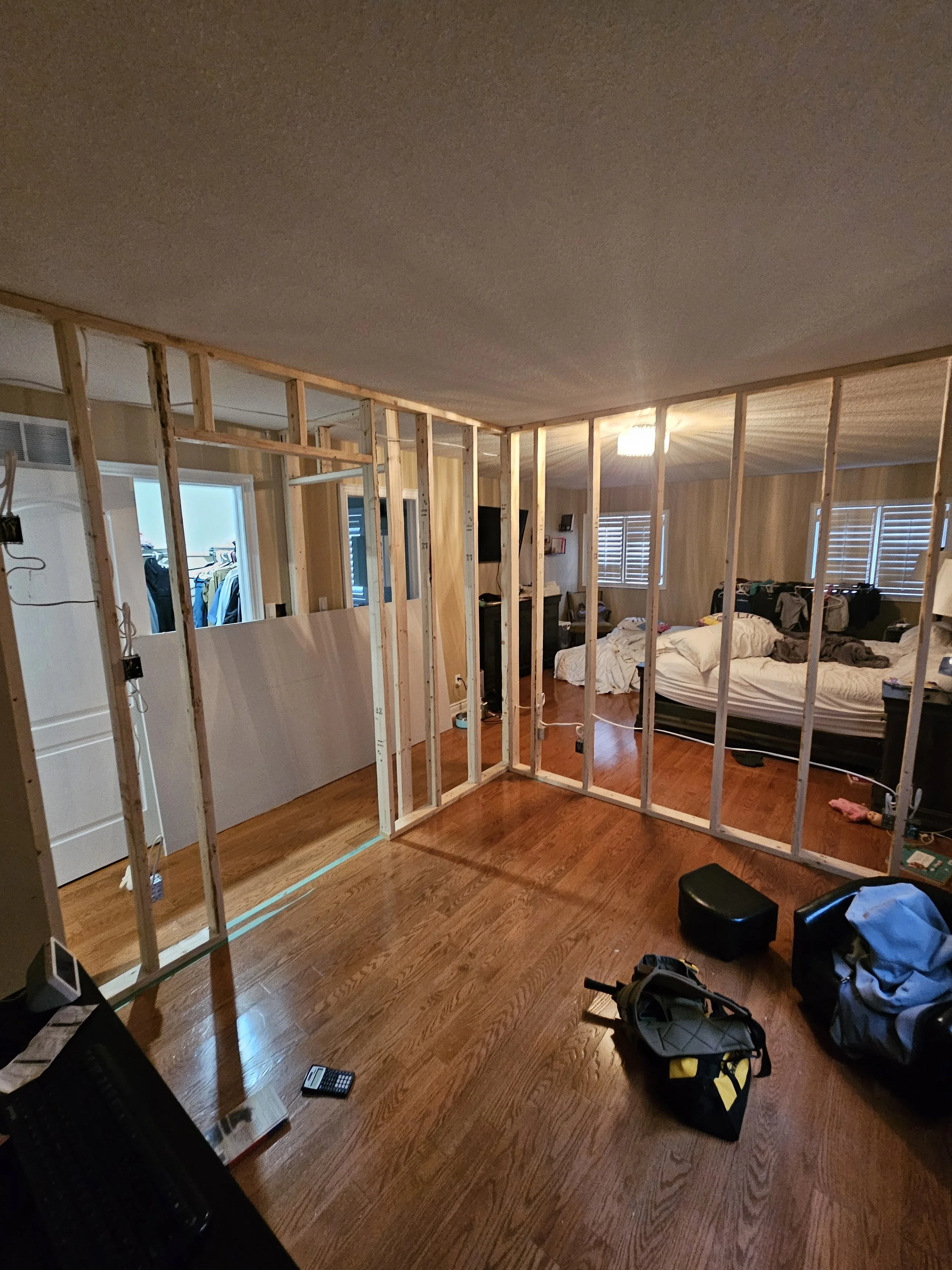 Room under renovation with exposed wooden framing, hardwood floors, and a bed in the background. Construction tools and bags are on the floor.