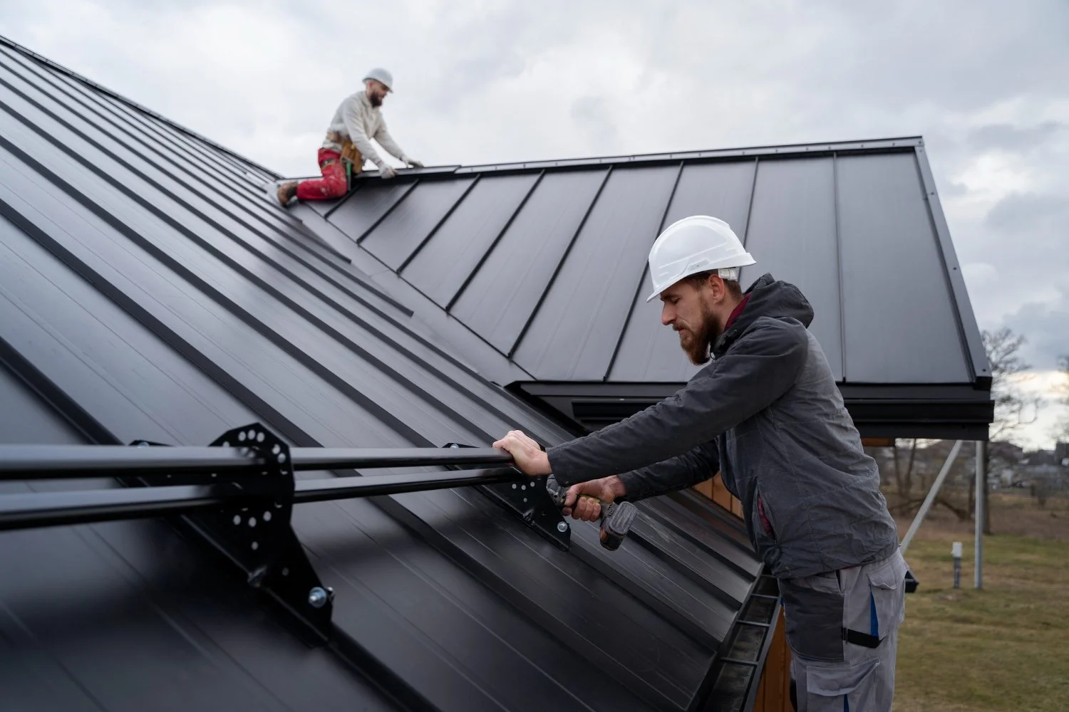 Two men installing solar panels on a metal roof. The man in the foreground is using a drill, and the man in the background is kneeling on the roof, securing the panels. Both are wearing safety helmets.