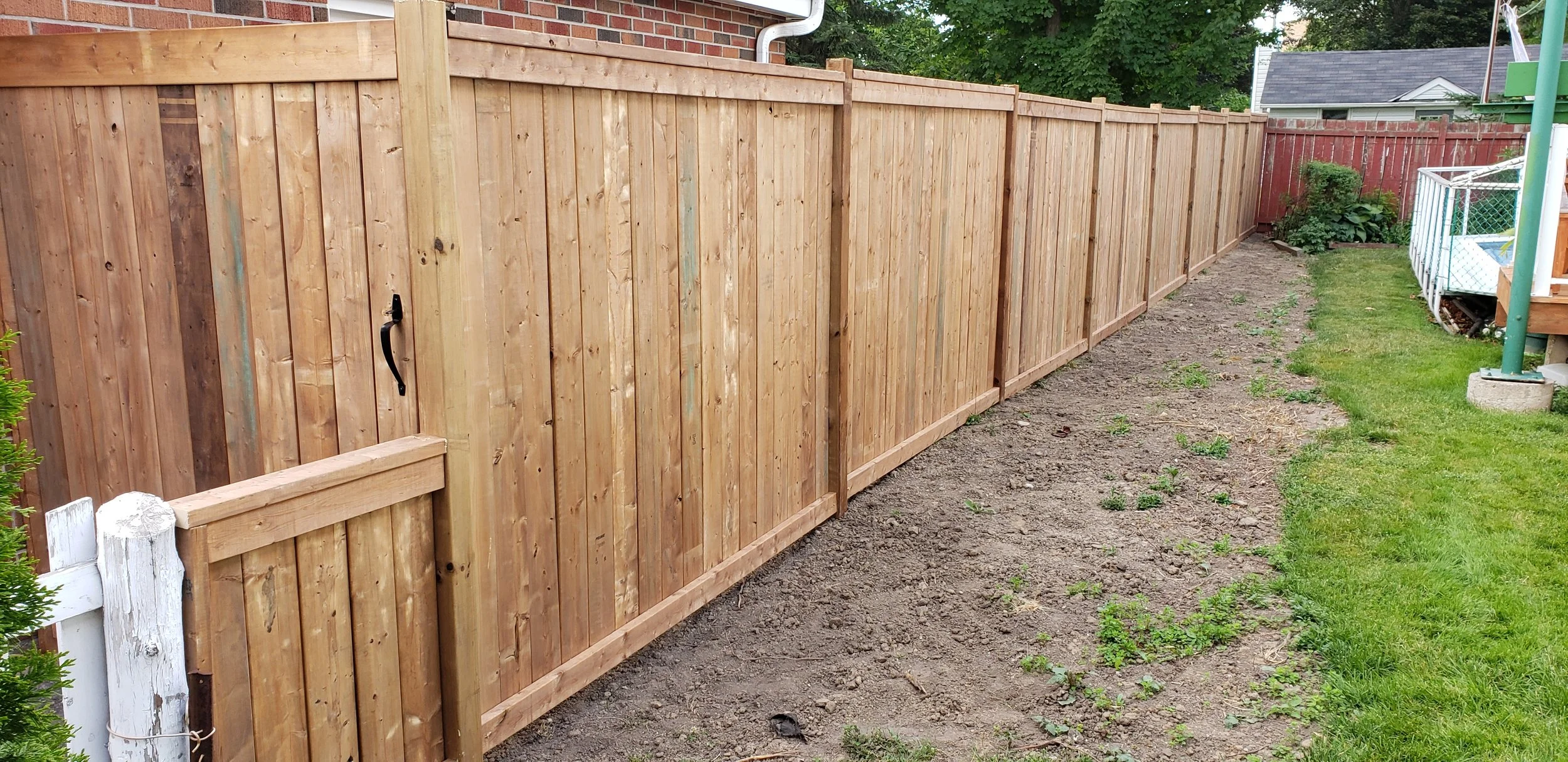 A backyard with a newly built wooden privacy fence along one side of the yard. The fence appears to be recently installed, with some dirt and small plants in front of it. On the right side, there's a grassy area and part of a deck or play area with a