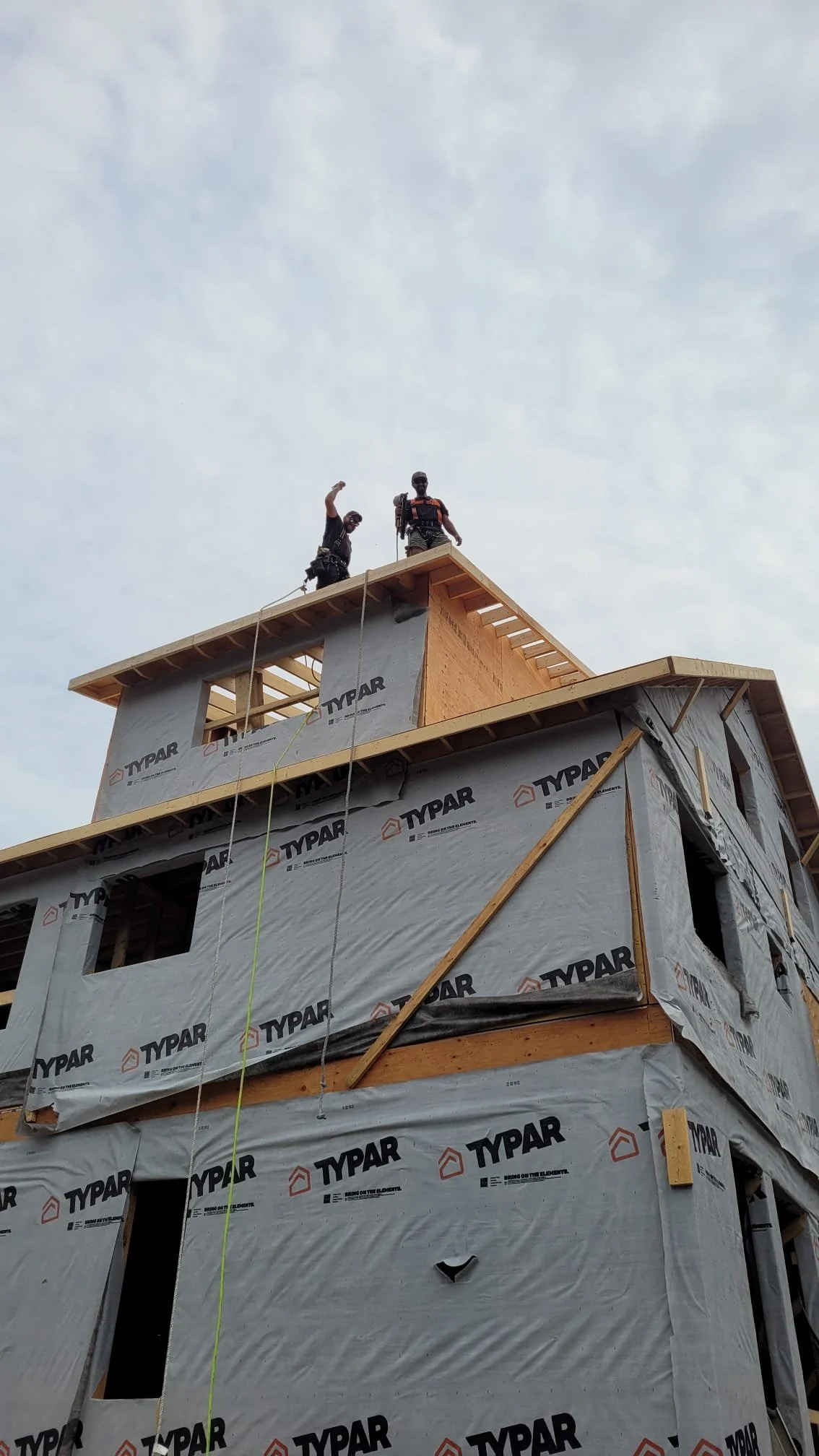 Two construction workers standing on the top of a multi-story building under construction, with one waving and the other standing with a toolbelt, against a cloudy sky.