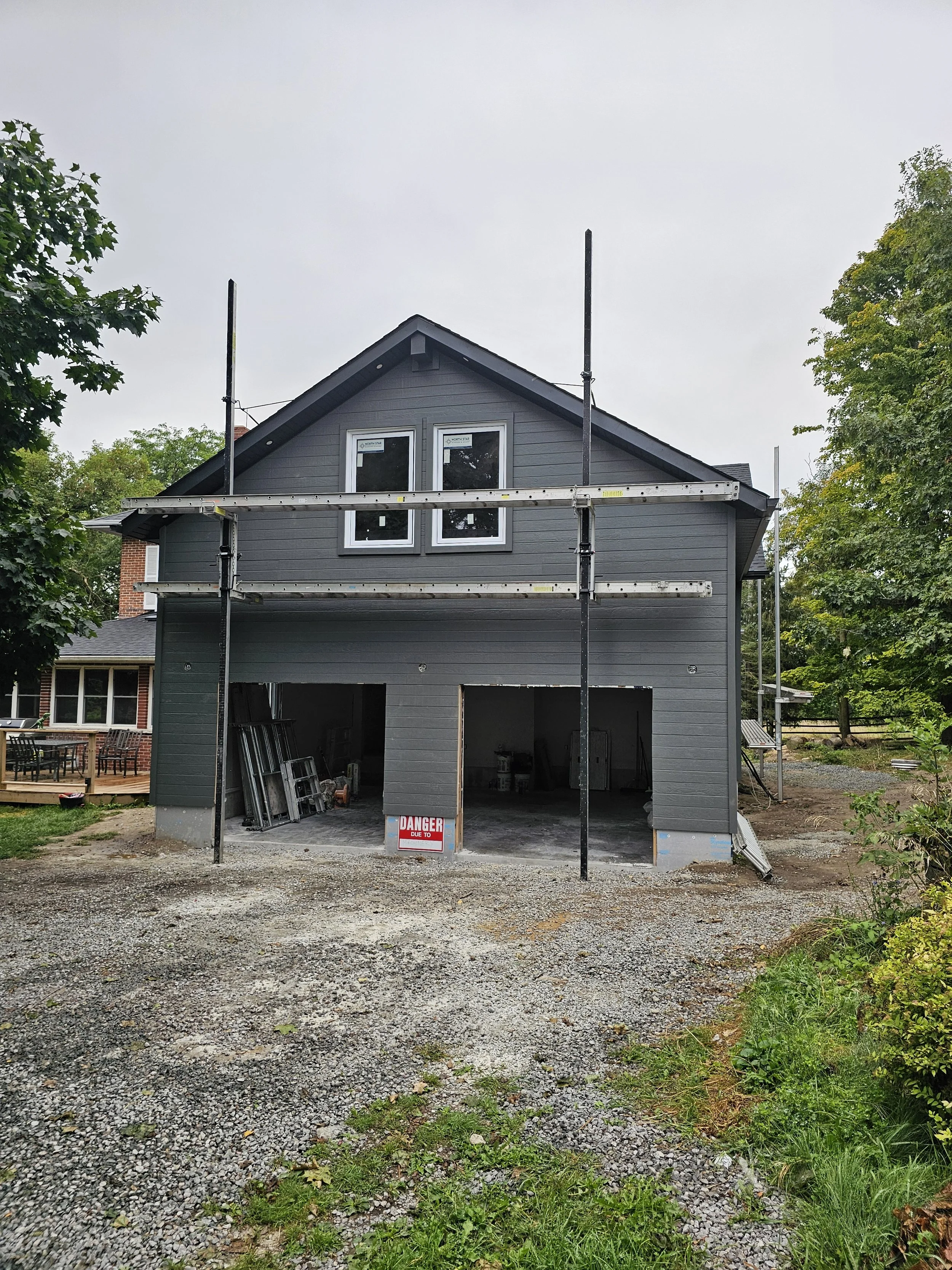 A two-story house under construction with dark gray siding, scaffolding around it, and two windows on the upper floor. The garage is open, showing construction materials inside, and a sign in front warns of danger.