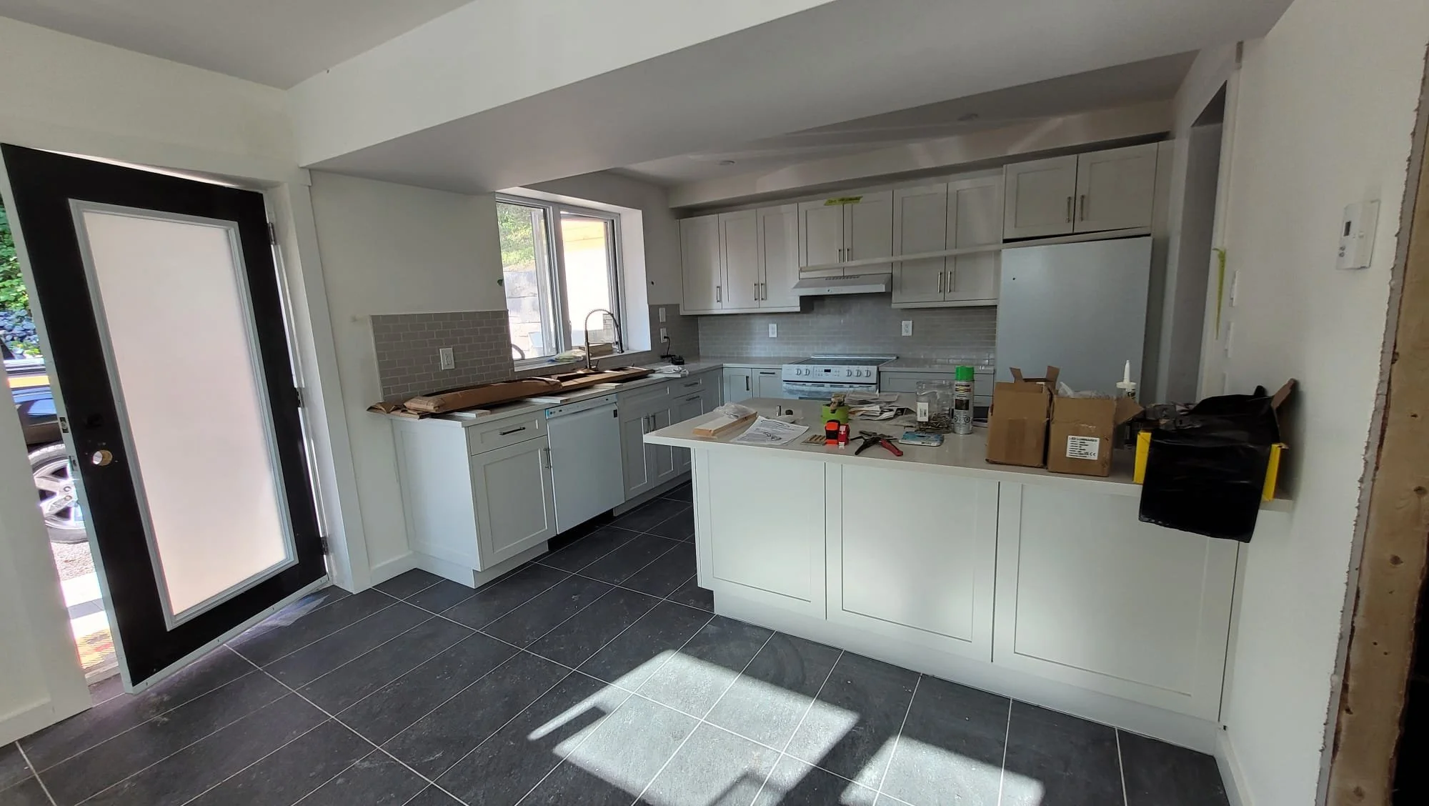 Kitchen under construction with white cabinets, gray countertops, gray backsplash tile, and black tile flooring. There are construction tools and materials on the counter.