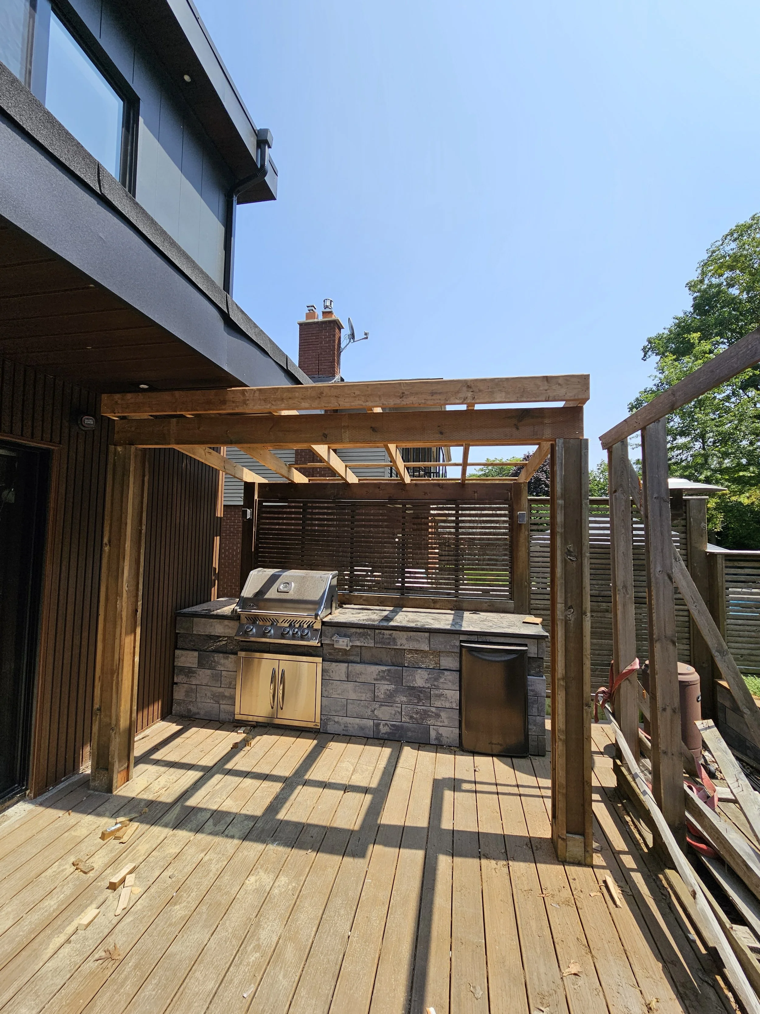Backyard deck with outdoor kitchen under construction, featuring a grill, small fridge, and wooden framing for a future roof or cover, with shadows cast by sunlight on the wooden floor.