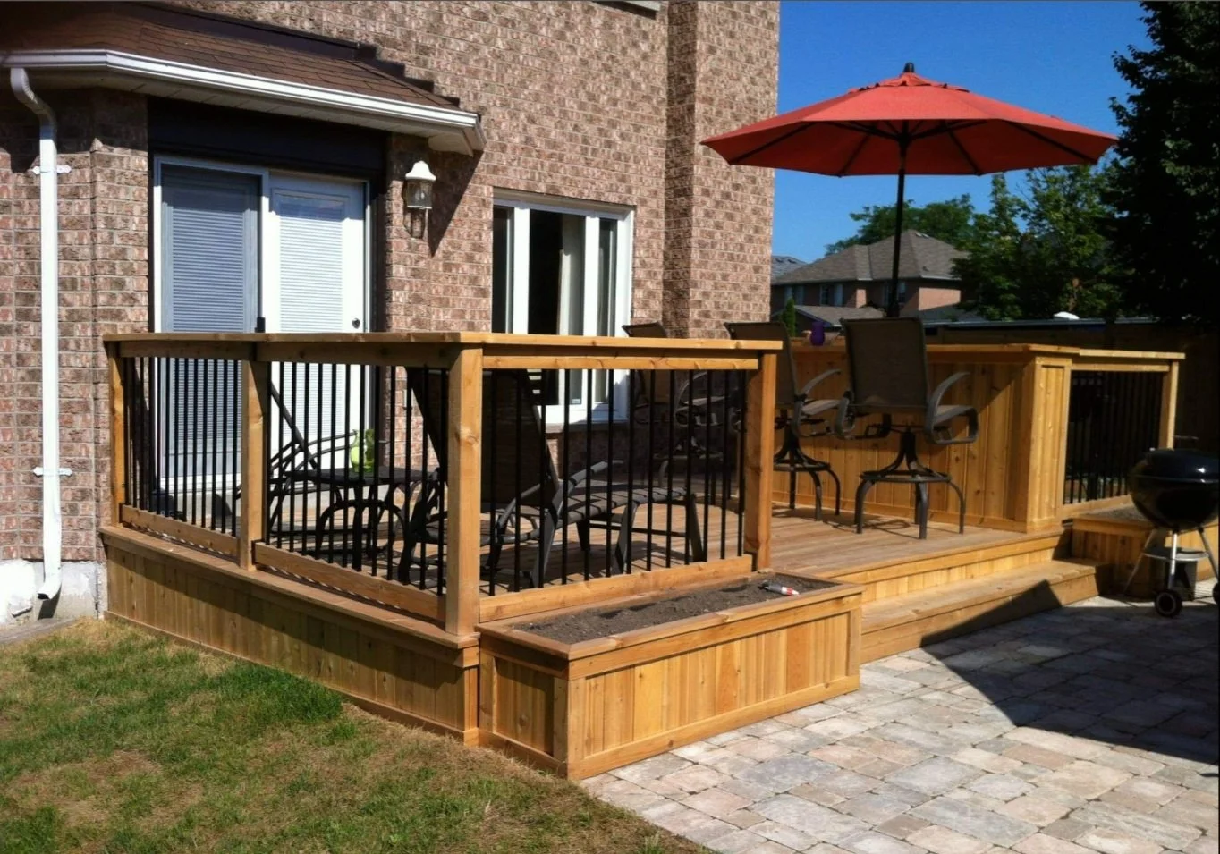 A wooden deck attached to a brick house with patio furniture, including a table, chairs, and a large red umbrella, overlooking a backyard with brick pavers and a grassy area.