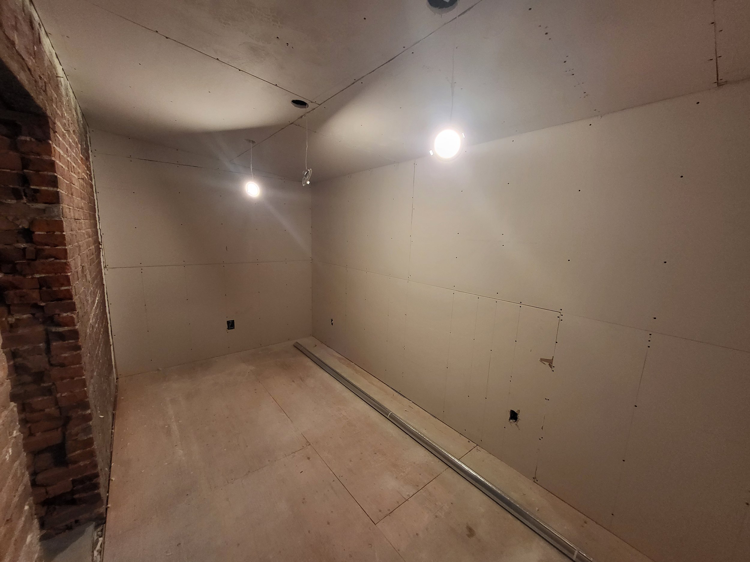 Empty room with unfinished walls, exposed brick on the left, drywall with visible screws, two ceiling lights, electrical outlets, and a metallic track on the floor.